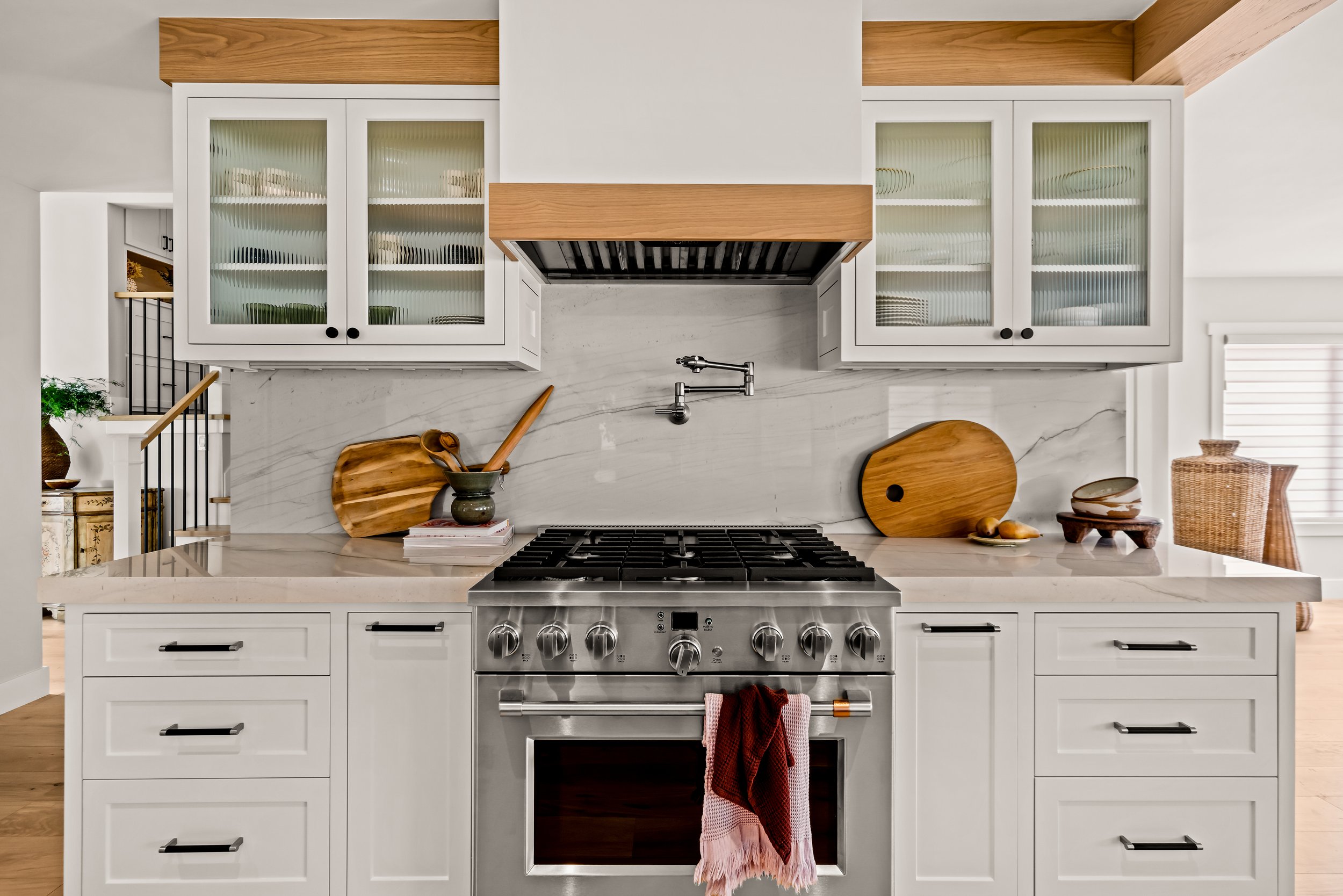 Modern white kitchen with a stainless steel stove, marble backsplash, and handcrafted wooden accents. Decorative cutting boards and dishware on the countertop.