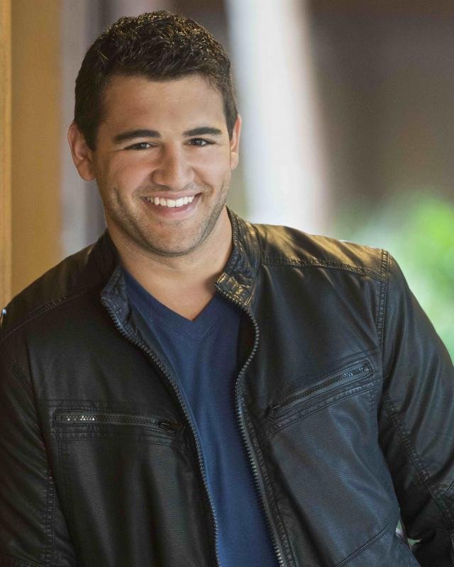 Shane Salanitri smiling, wearing a black leather jacket over a blue shirt, standing indoors near a window.