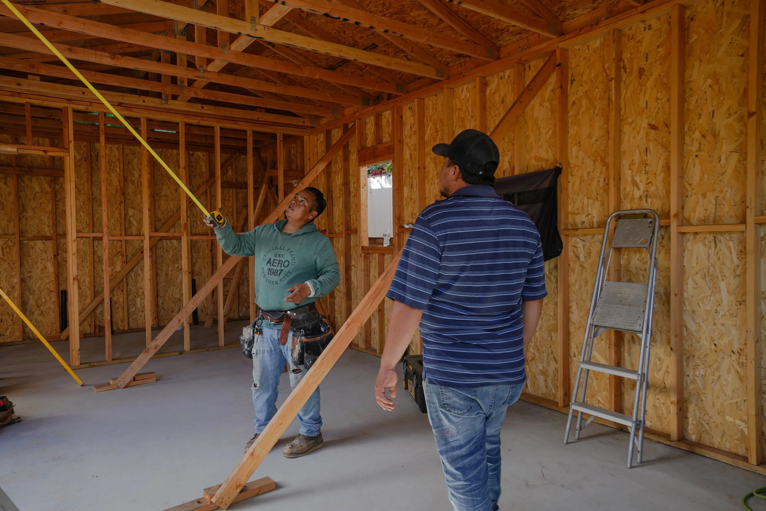 Two men working inside a house under construction in San Diego, with exposed wooden framing. One man is holding a measuring tape and showing it to the other man. There is a ladder leaning against the wall behind them.