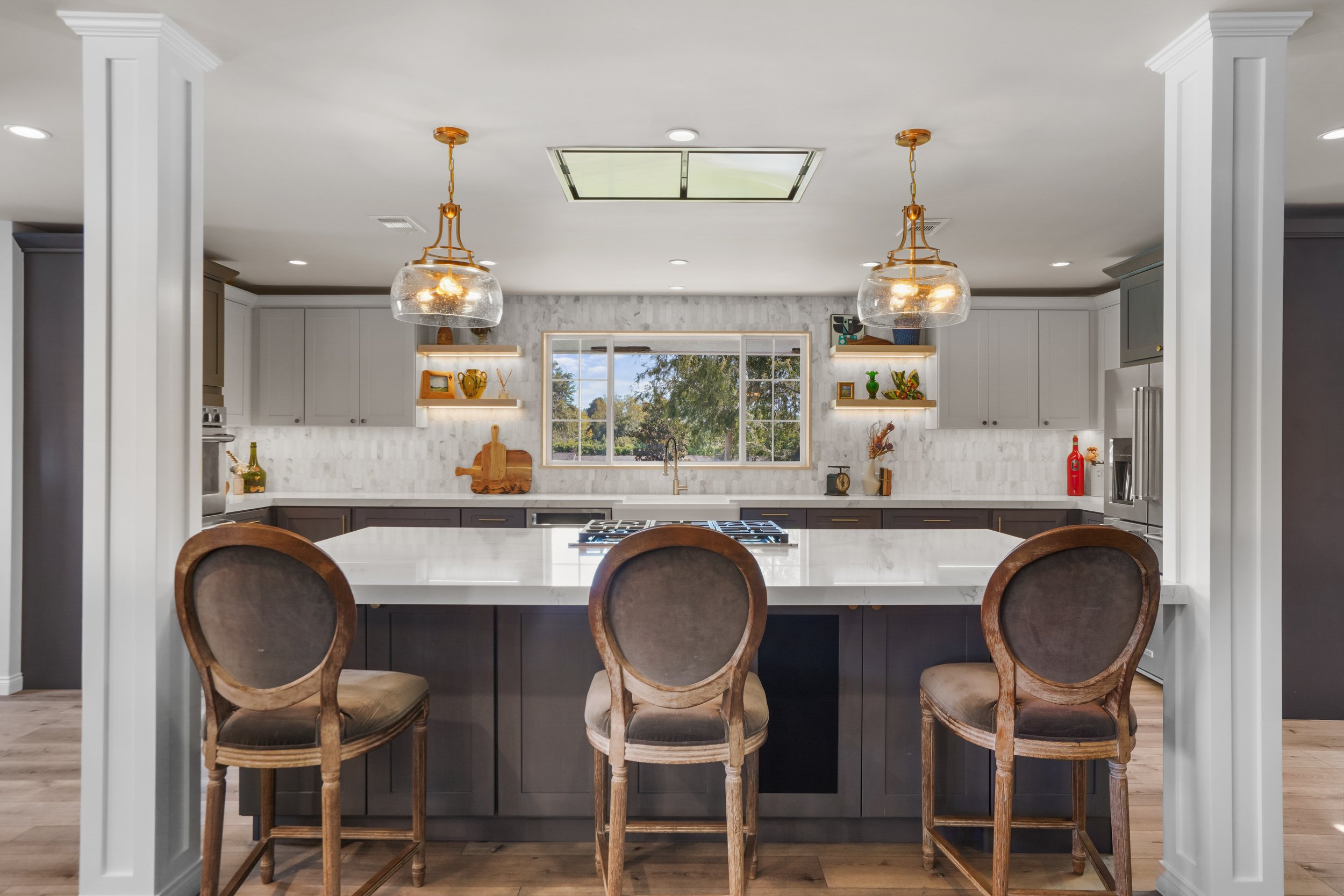 Modern kitchen with white cabinets, a large white marble island, three wooden chairs, and a window overlooking trees. Two glass pendant lights hang above the island, and shelves with decorative items are on the wall.