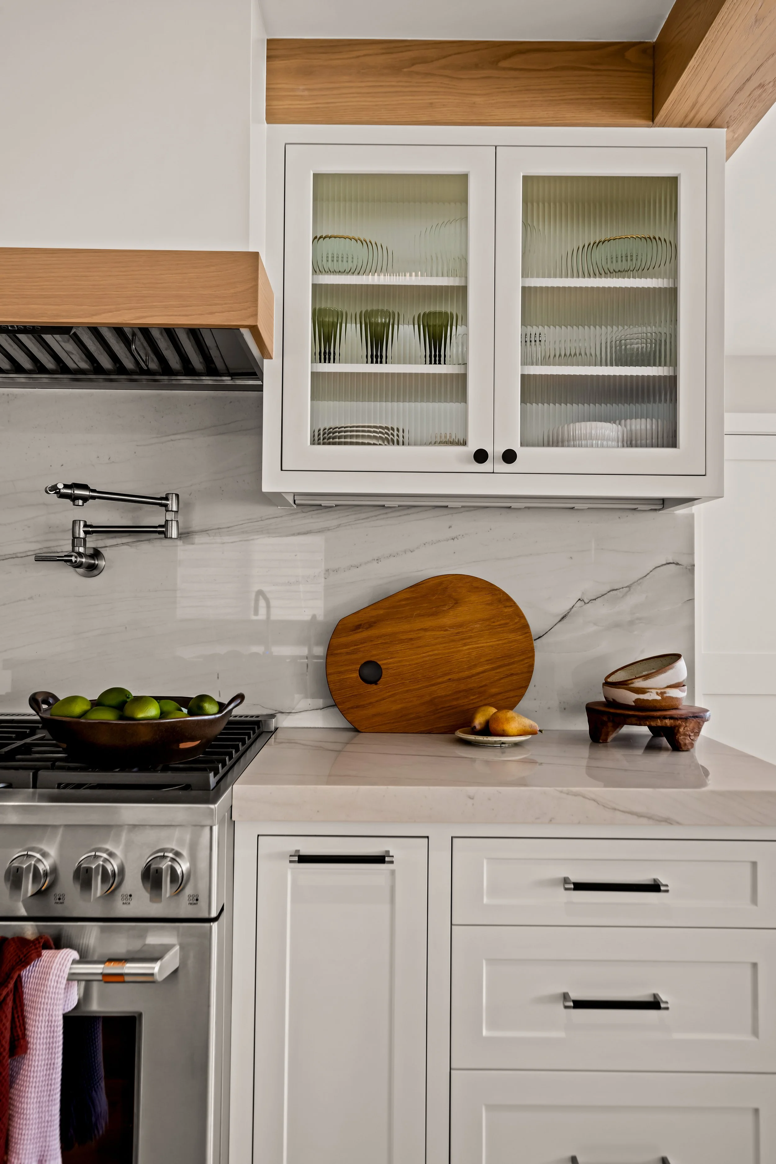 Modern Orange County kitchen with white cabinets, a marble countertop, a stainless steel stove, a glass-front cabinet filled with dishes, a wooden cutting board, a pottery bowl, limes, pears, and a decorative bowl.