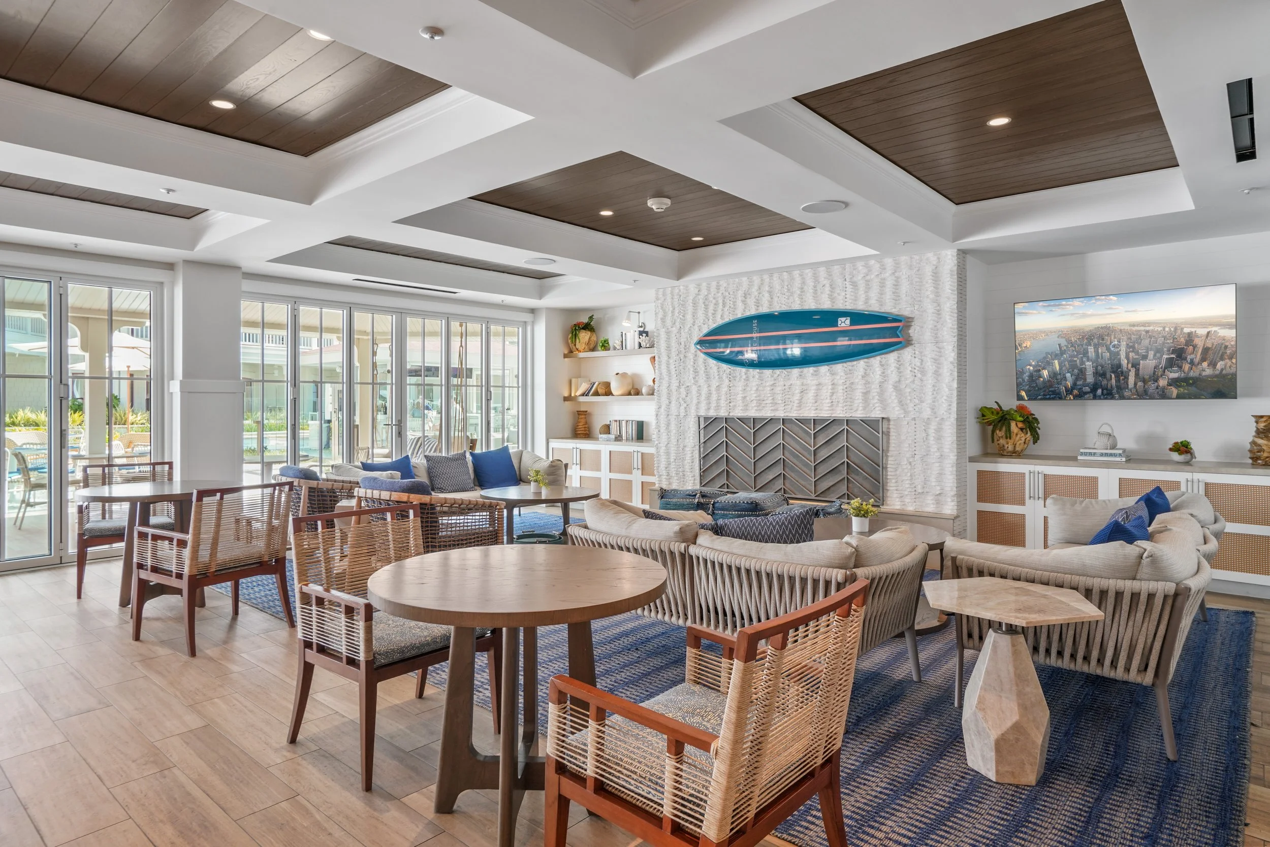 Bright and airy living room at Hotel Del Coronado with sliding glass doors, wooden ceiling accents, sitting area with sofas and chairs, a wall-mounted TV, and a surfboard decoration.