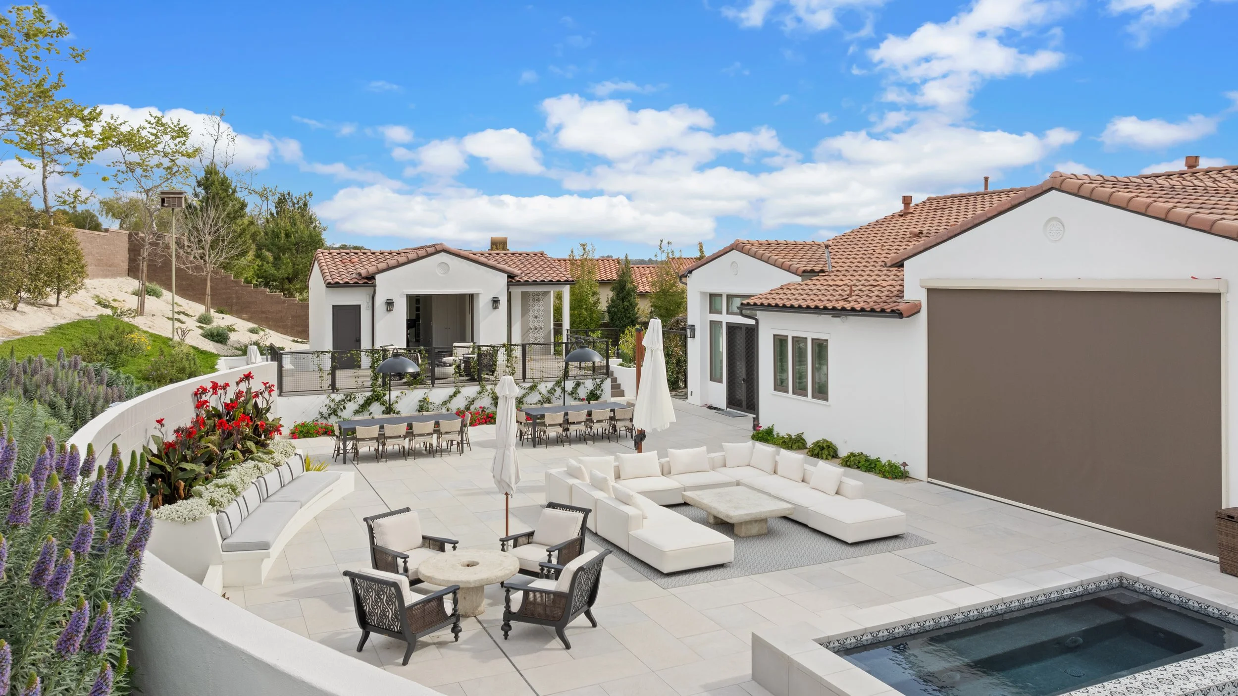 Luxury resort-style backyard remodel in San Diego with a seating area, umbrella-shaded dining tables, a hot tub, and landscaped garden beds, surrounded by white stucco houses with red tile roofs under a blue sky with clouds.