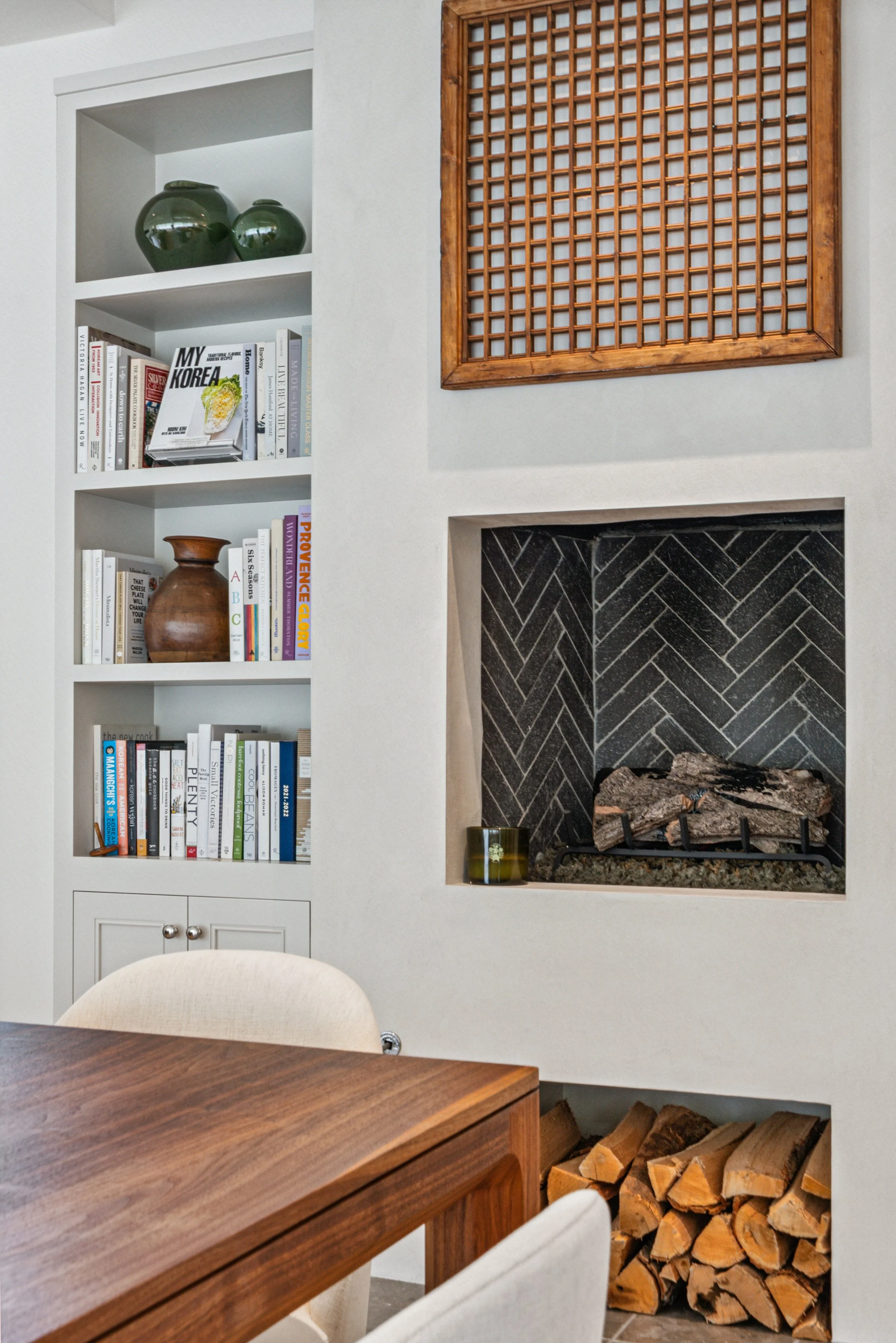 Interior of a living room corner featuring a built-in white bookshelf with books and decorative vases, a fireplace with dark brick surround and a wood pile underneath, and a wooden wall panel above the fireplace.