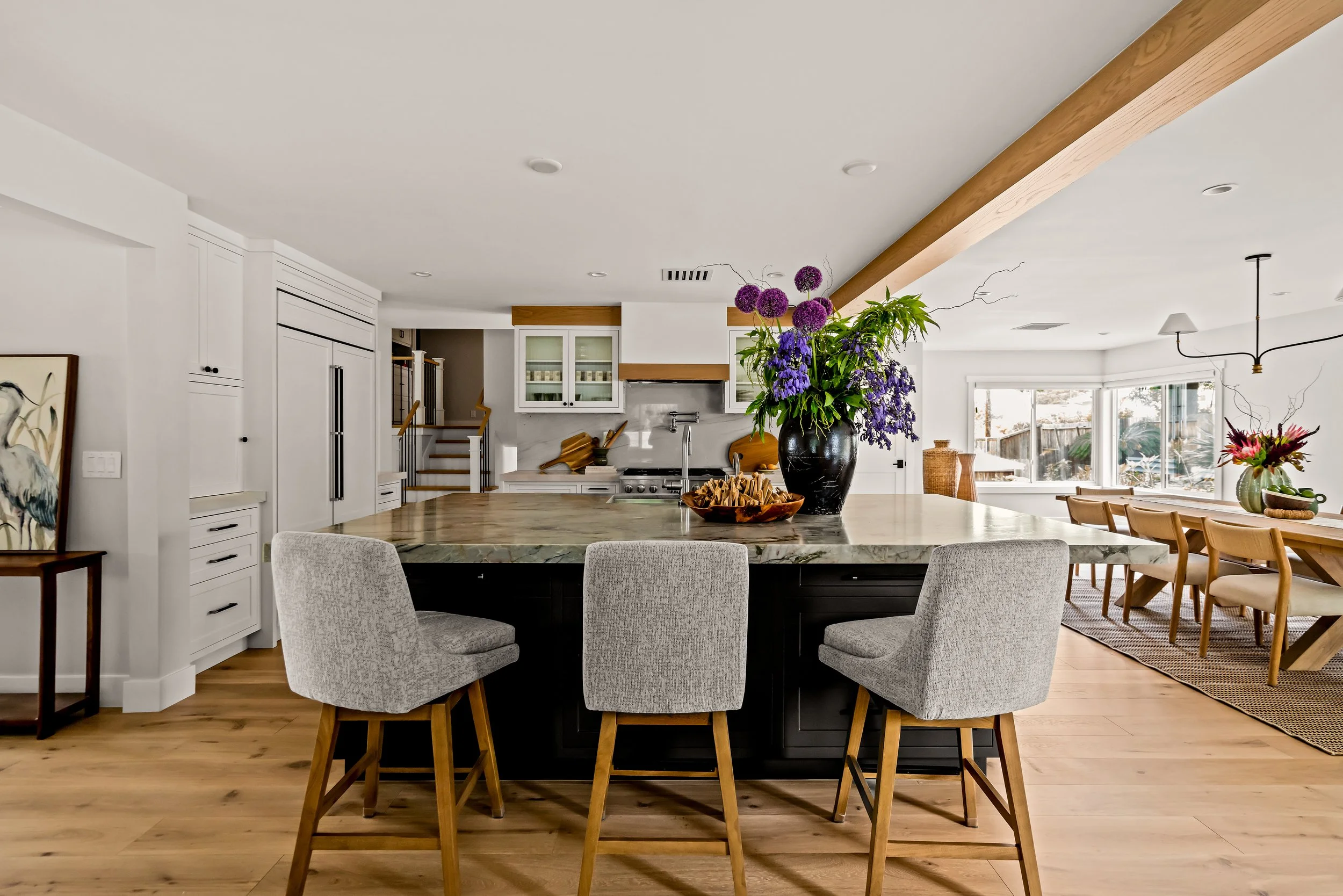Modern kitchen with a large island, gray chairs, and a black vase with purple flowers, adjacent to a dining area in a bright, open space.