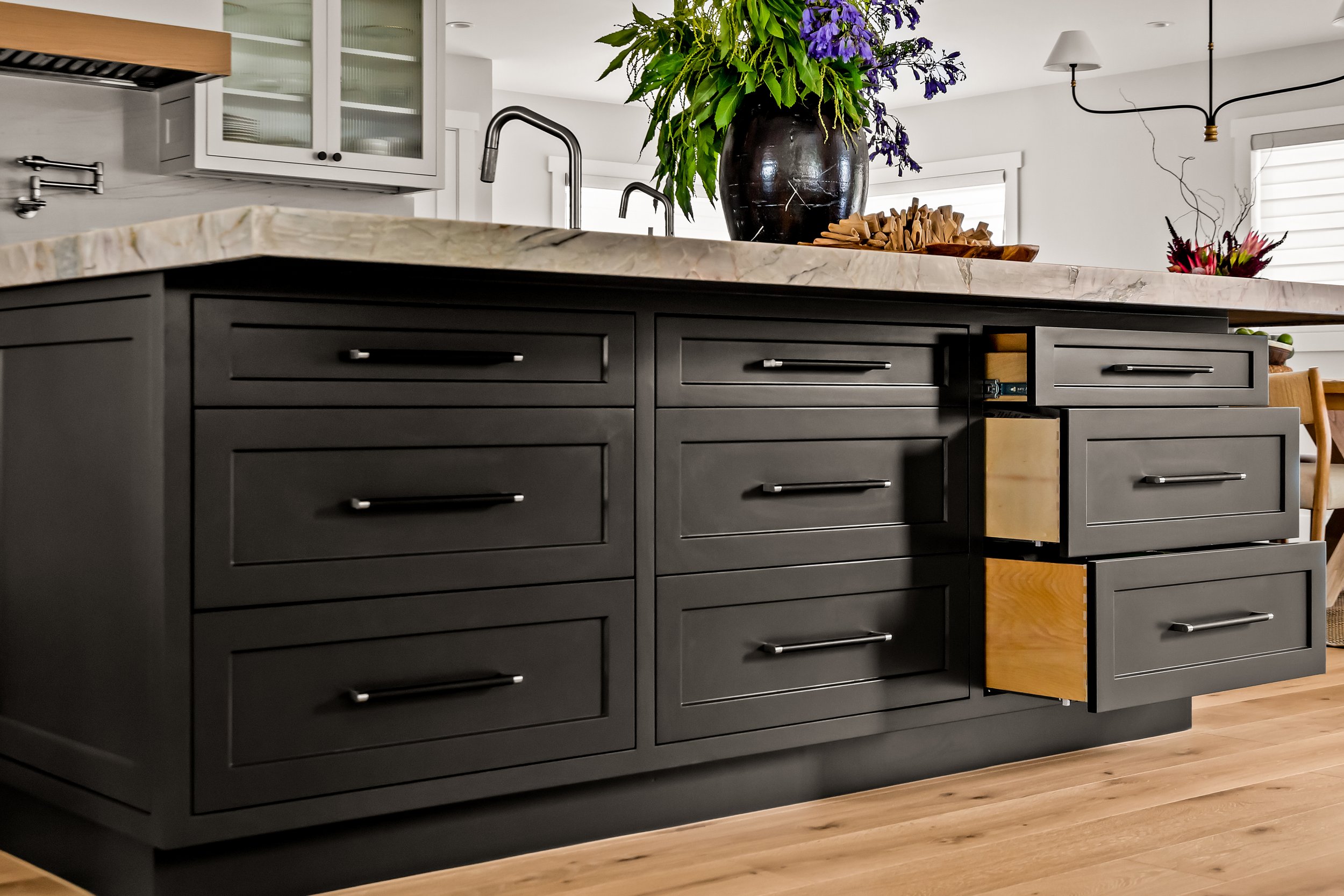 Kitchen island with black drawers, marble countertop, and a large black vase with purple and green flowers on top.