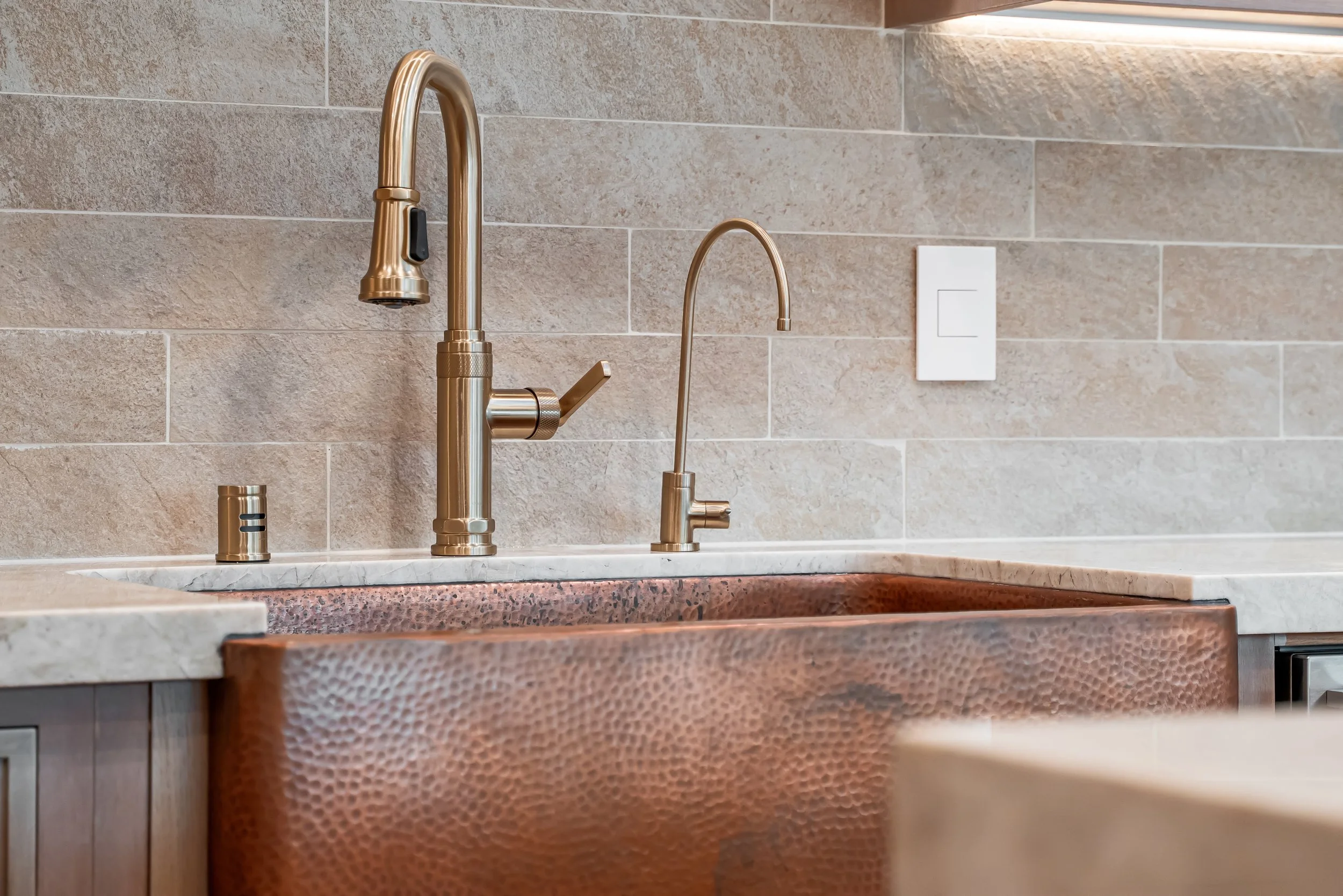 Kitchen sink area with a copper hammered finish sink, gold-tone faucets, and a beige ceramic tile backsplash.