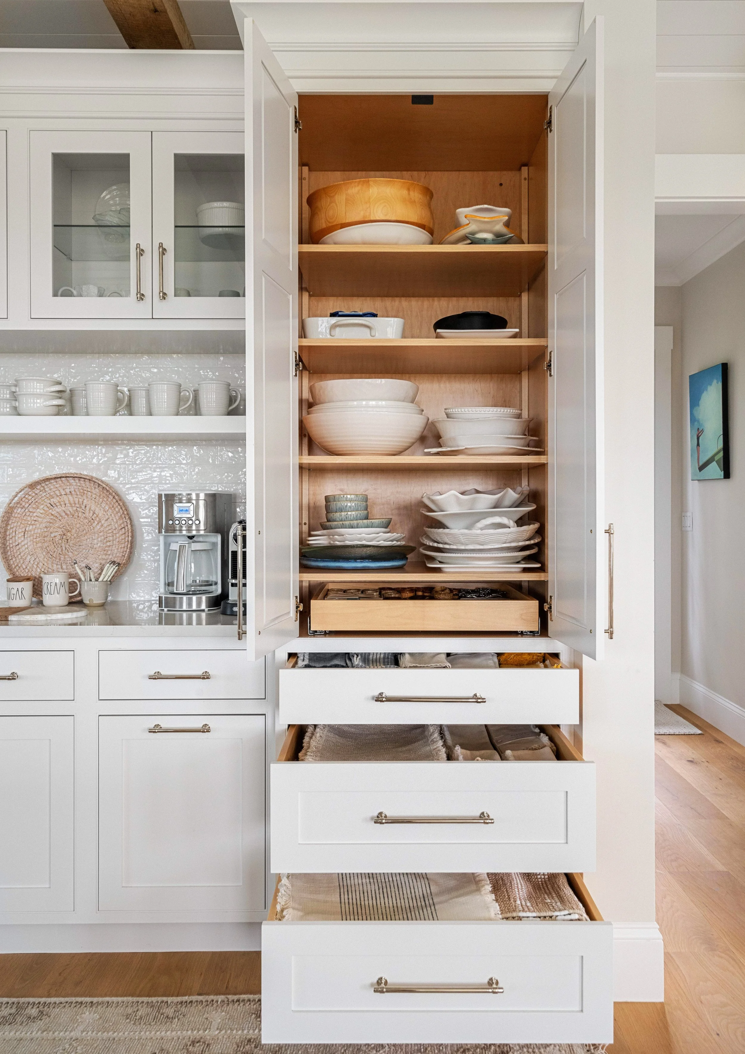 Open kitchen cabinet with various dishes, bowls, and plates arranged on shelves, adjacent to a countertop with mugs, a coffee maker, and a small tray with cups.
