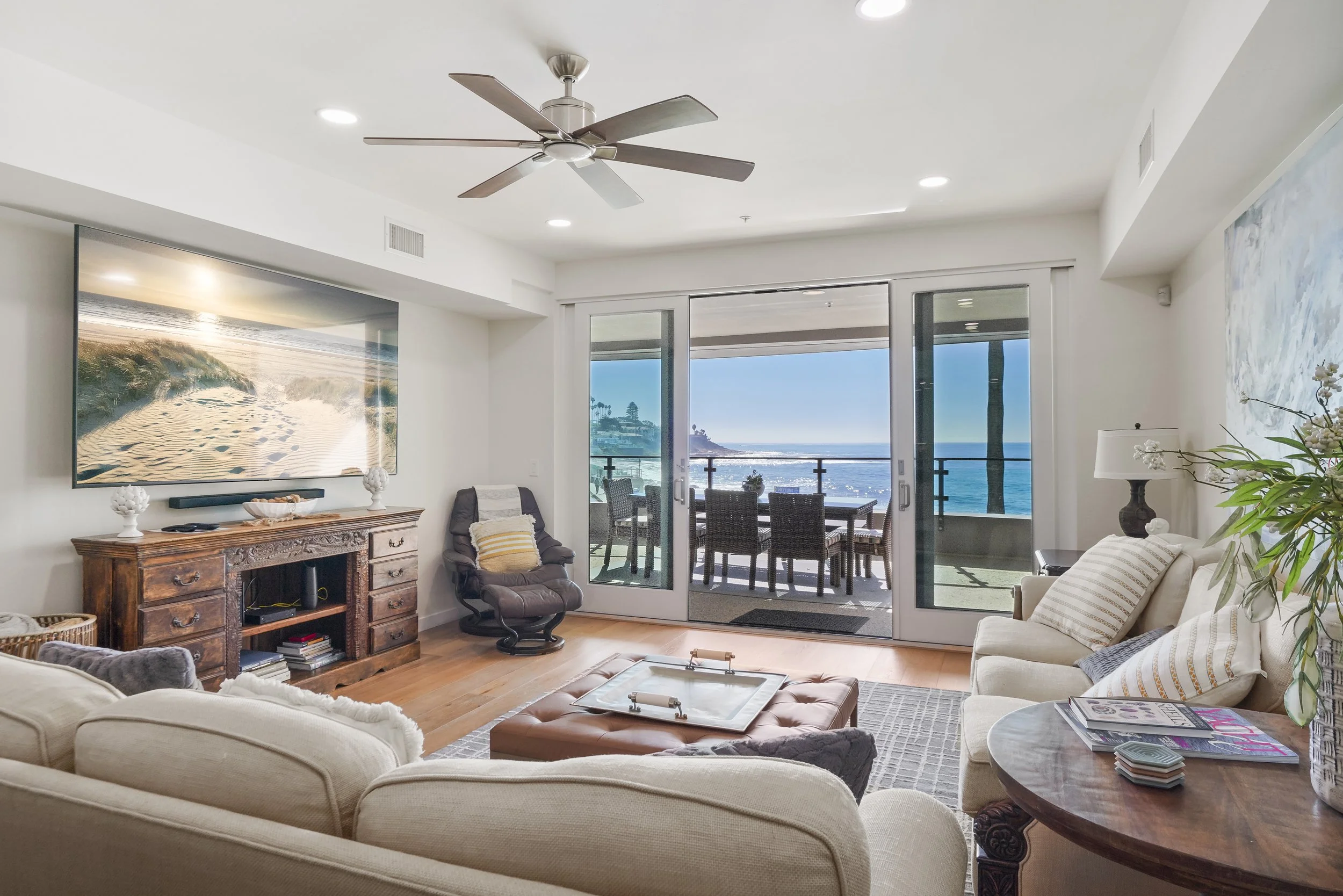 Living room with La Jolla ocean view through sliding glass doors, beige sofas, a wooden coffee table, a large wall-mounted TV, a ceiling fan, and outdoor balcony with dining table and chairs.
