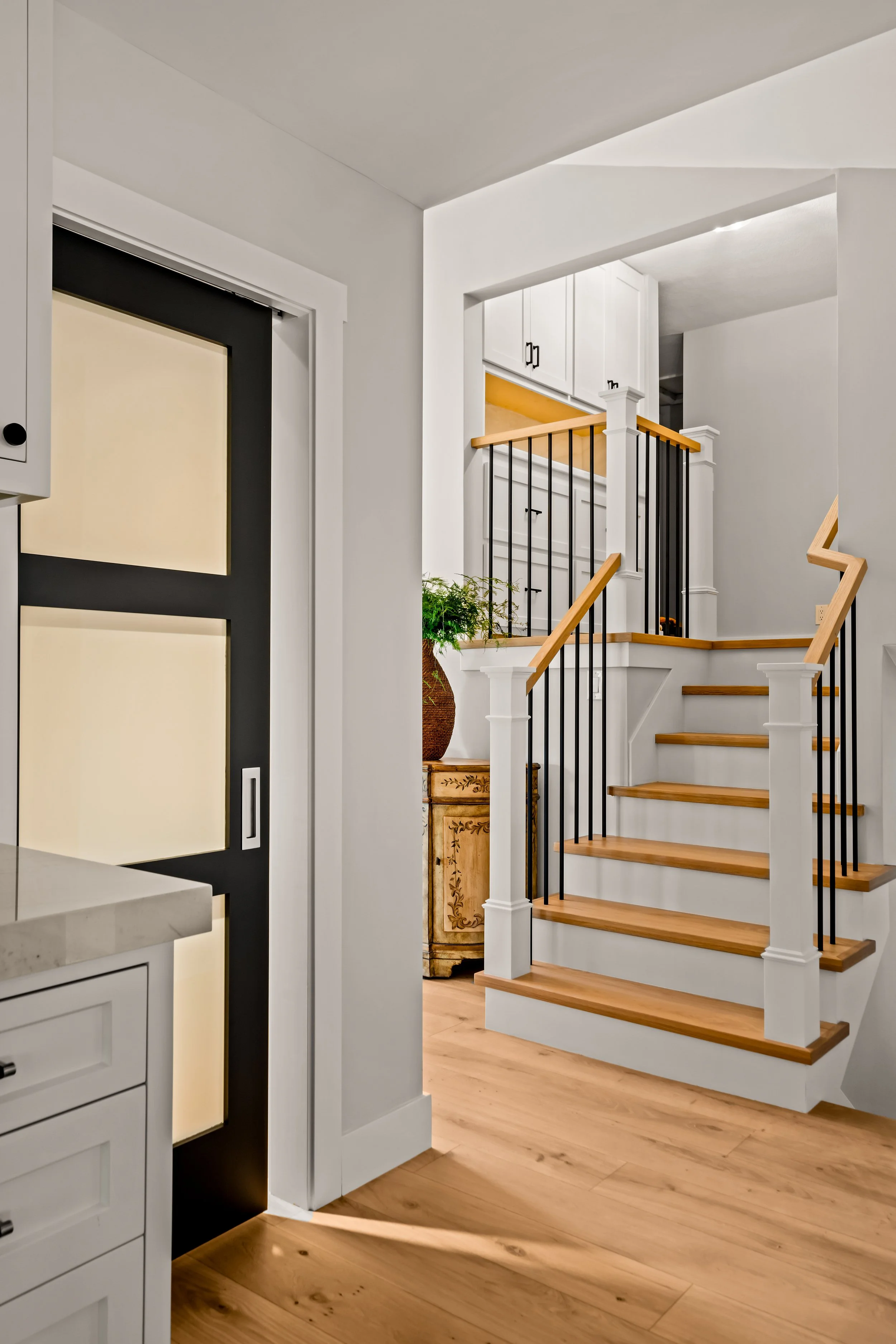 Interior view of a modern coastal home with a staircase featuring wooden steps and white risers. The home includes white and beige custom cabinetry, a marble countertop, and a potted plant, with natural wooden flooring.