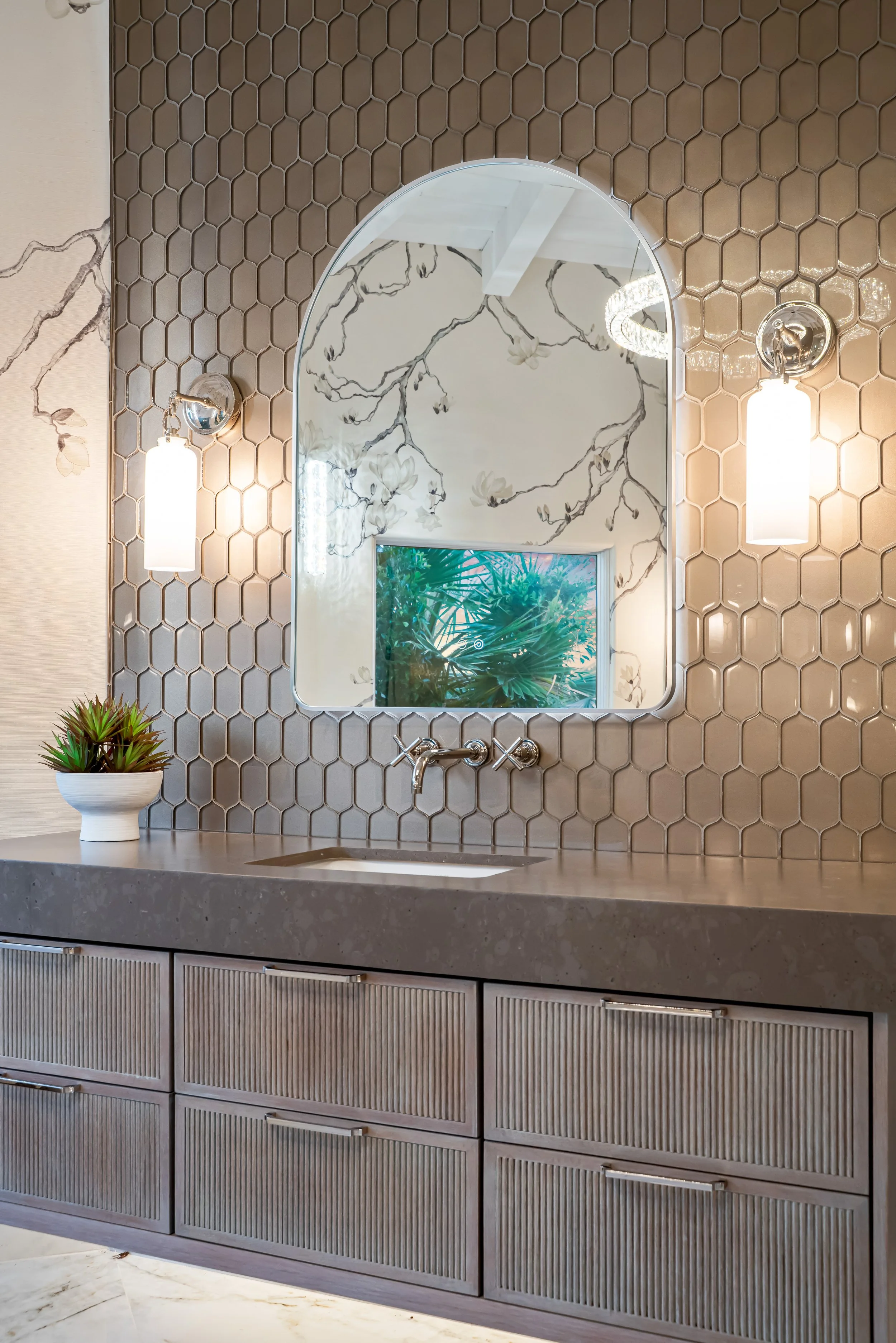 Modern bathroom vanity with a gray countertop, three drawers, and a small sink. Above is an arched mirror with a honeycomb tile backsplash, flanked by two wall-mounted light fixtures. A window with greenery outside is reflected in the mirror.