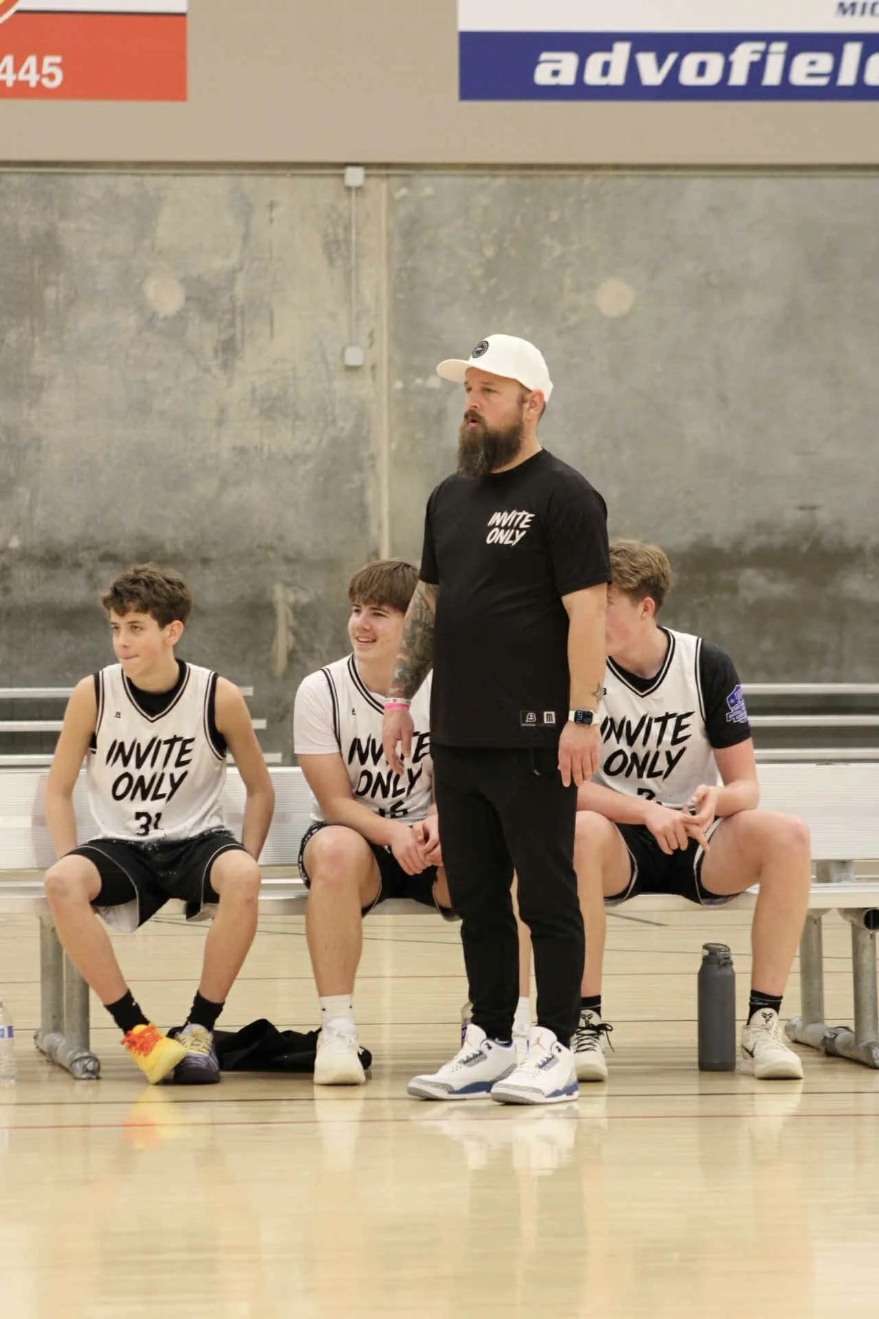 A basketball coach standing in front of three players sitting on a bench during a game, all wearing white jerseys with 'INVITE ONLY' printed on them. The coach is wearing a black t-shirt, black pants, and a white cap. The environment is an indoor basketball court.