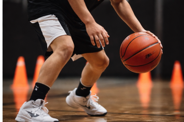 Basketball player practicing dribbling on indoor court with orange cones in the background.