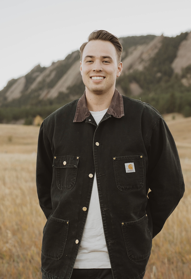 A young man wearing a black Carhartt jacket standing in an open field with mountains in the background, smiling at the camera.