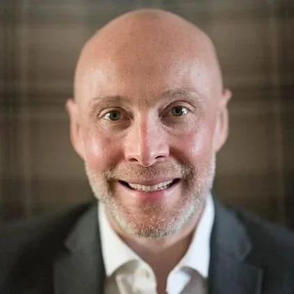 Man with a shaved head, beard, and mustache, smiling, wearing a suit jacket and white shirt, indoor setting with wood-paneled background.