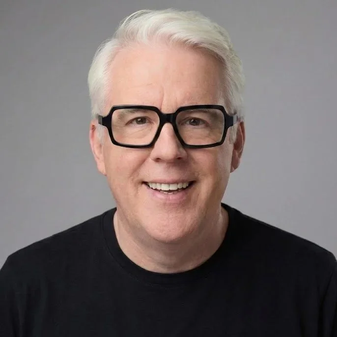 A middle-aged man with white hair, black glasses, and a black shirt smiling at the camera against a plain gray background.