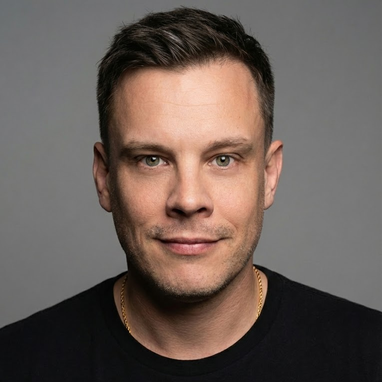 Close-up portrait of a man with short dark hair, fair skin, and blue eyes, wearing a black shirt and a gold chain against a gray background.