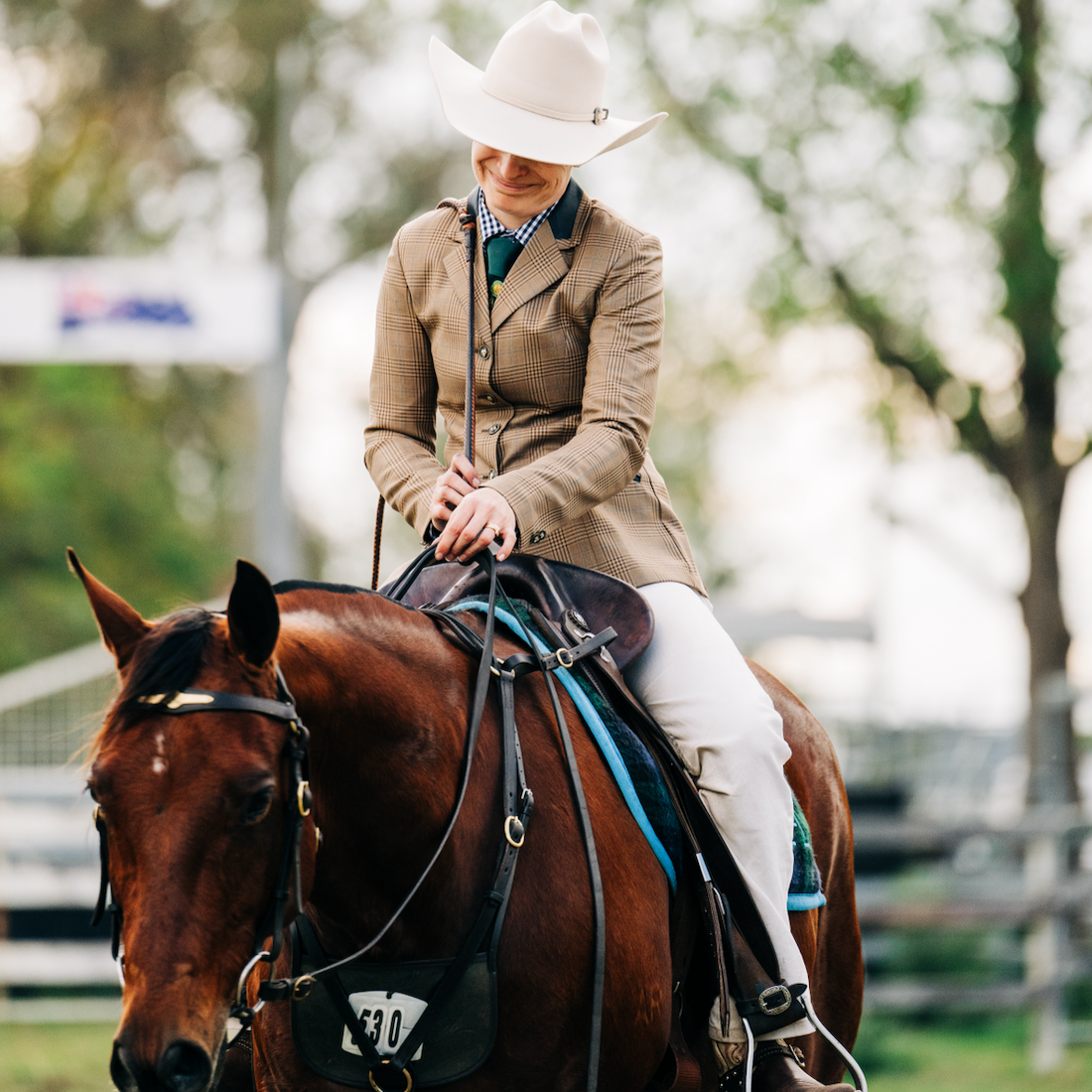 A smiling woman wearing a white cowboy hat and a checkered blazer riding a brown horse during an outdoor event.