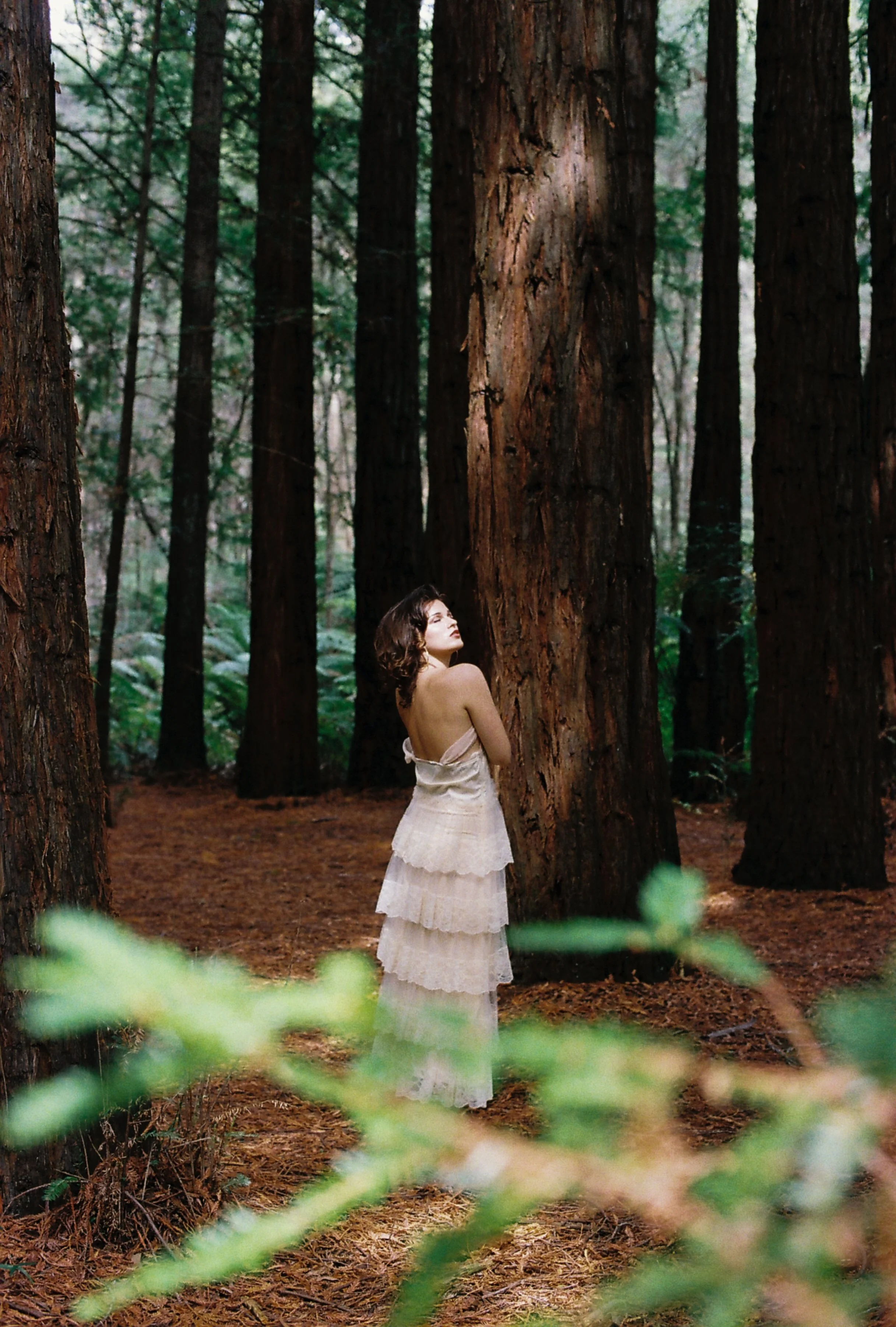 A woman in a white, layered dress standing in a dense forest with tall trees and green foliage.