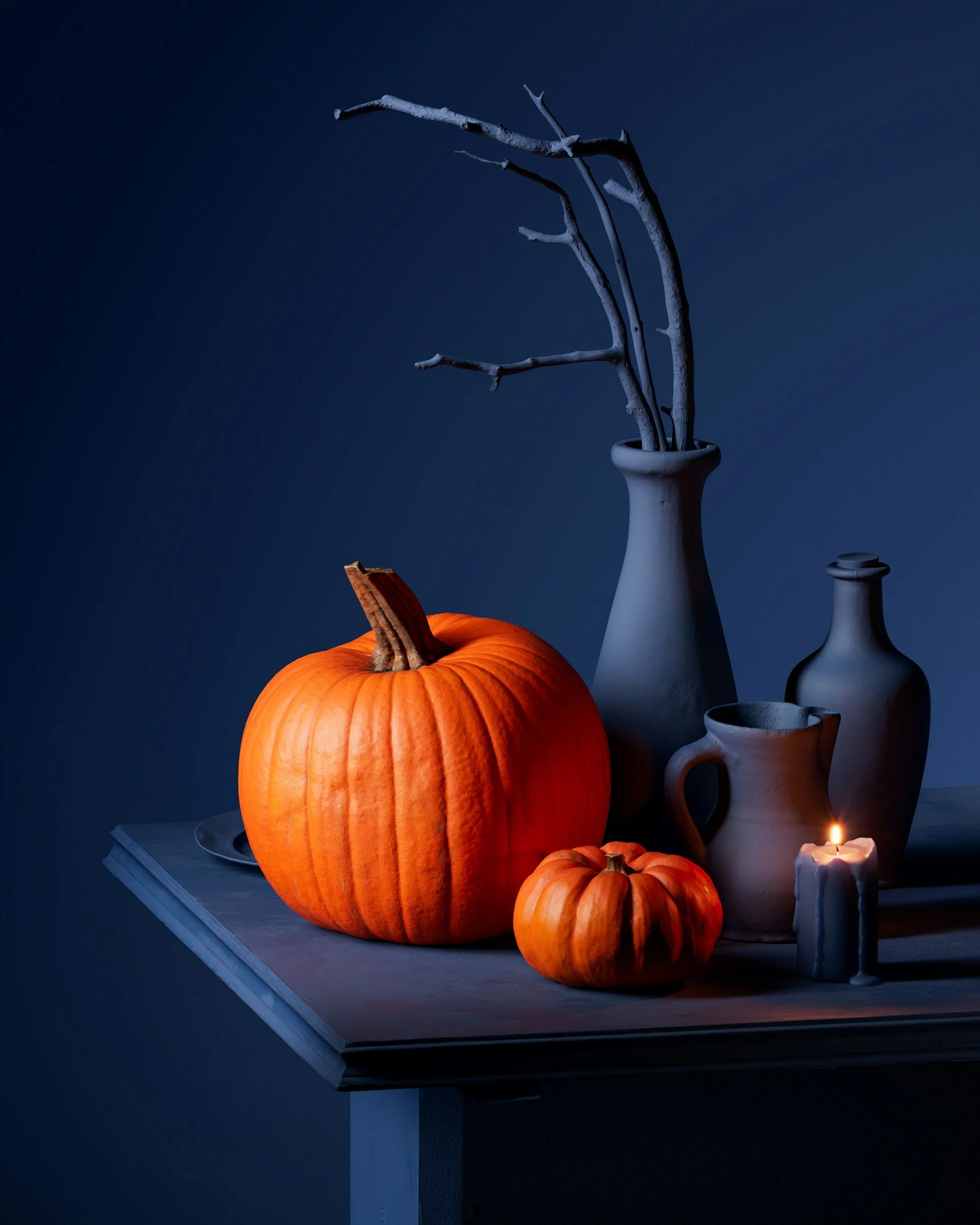 An orange pumpkin and a smaller pumpkin on a wooden table, with ceramic vases and a lit candle, set against a dark background.