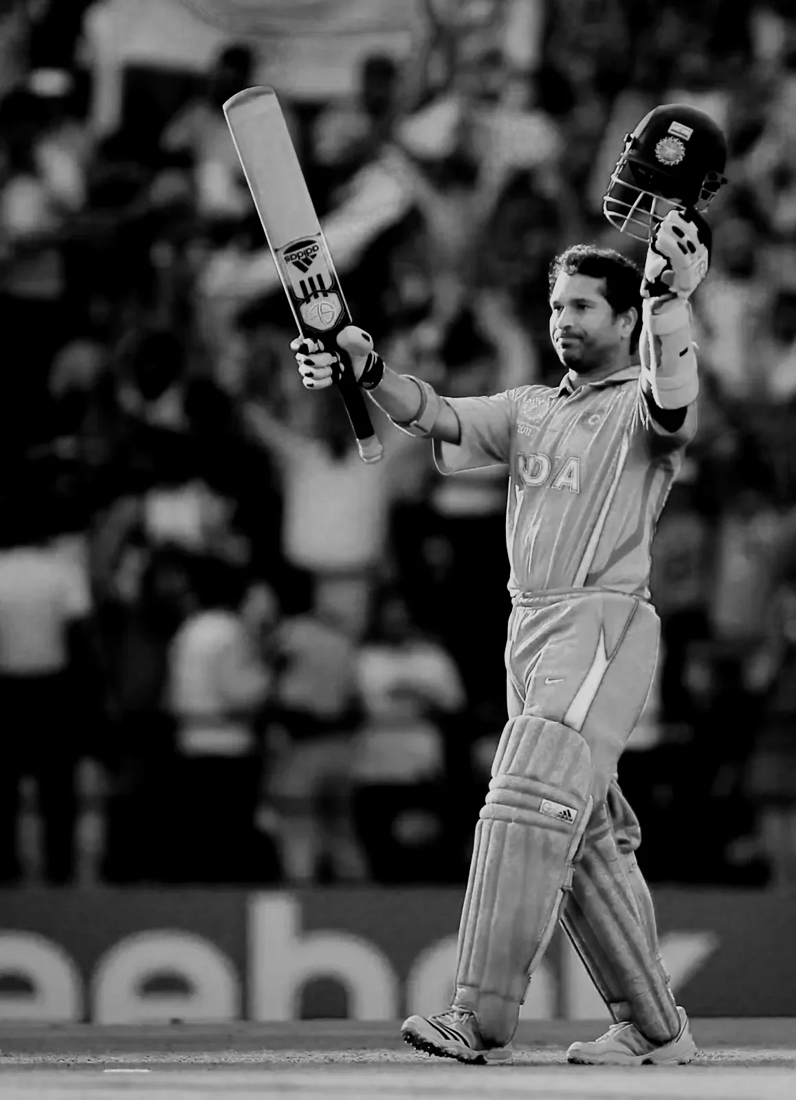 A cricket player in protective gear holding a helmet and bat, celebrating on the cricket field.