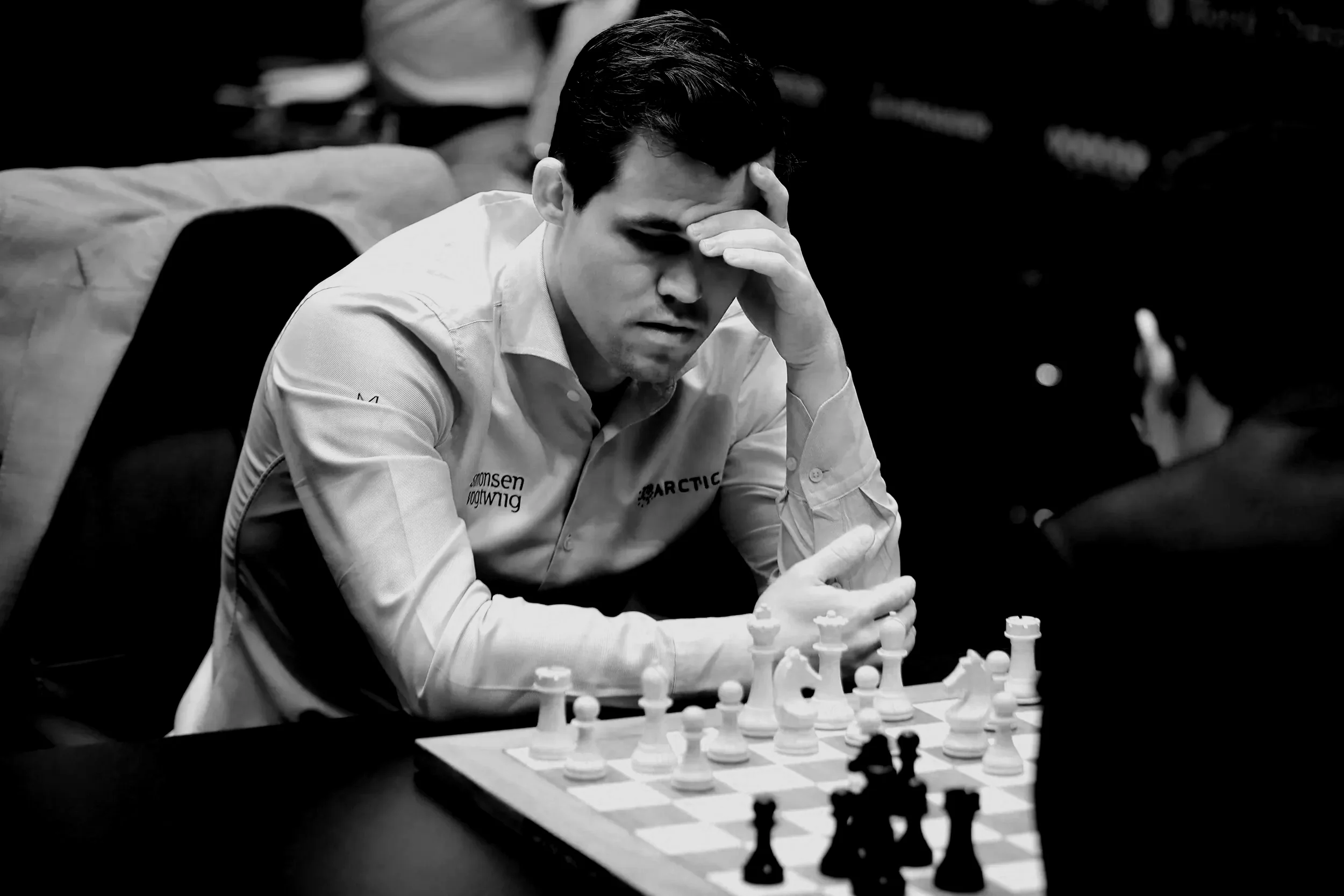 Magnus Carlsen wearing a white shirt, sits at a chessboard contemplating his move during a chess match. His hand supports his forehead as he focuses on the game.