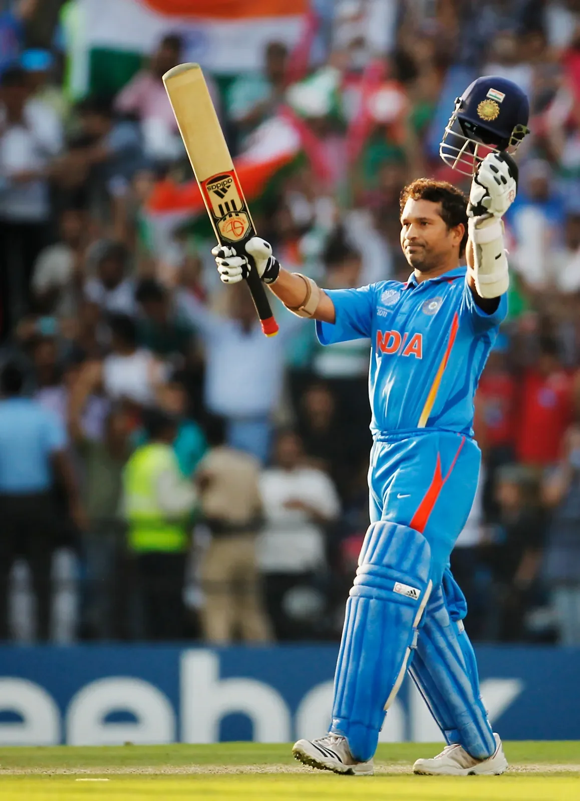 An Indian cricket player wearing blue uniform holding a cricket bat and a helmet, celebrating on the field with a crowd in the background.