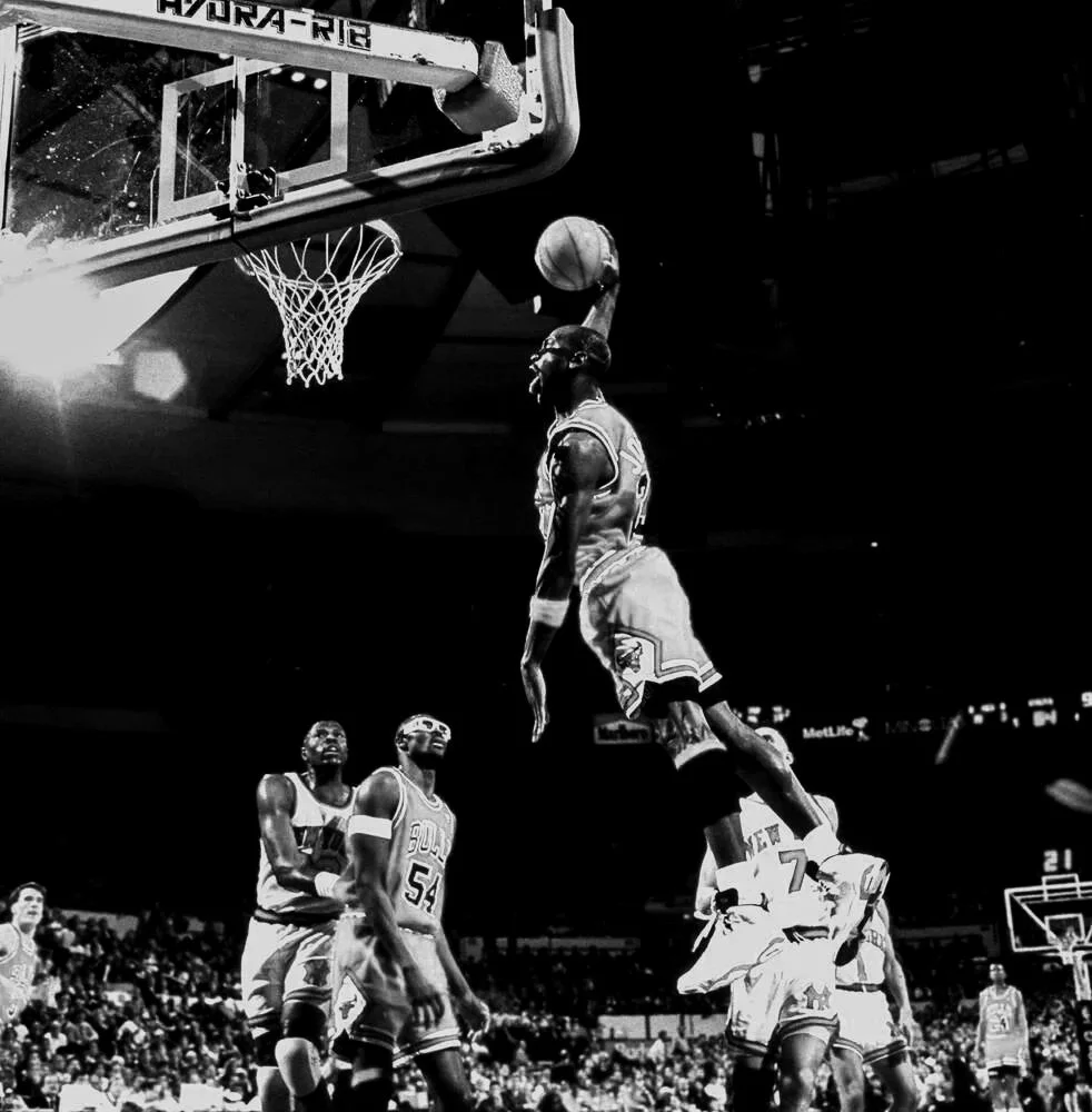 Michael Jordan mid-air executing a slam dunk at the basketball hoop during a game in a packed arena.
HNDRX AI