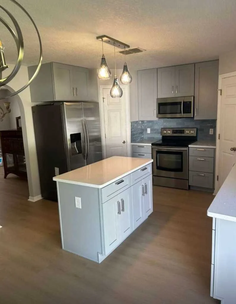 Modern kitchen with gray cabinets, stainless steel refrigerator and oven, gray tiled backsplash, white kitchen island, and hanging pendant lights over the island.