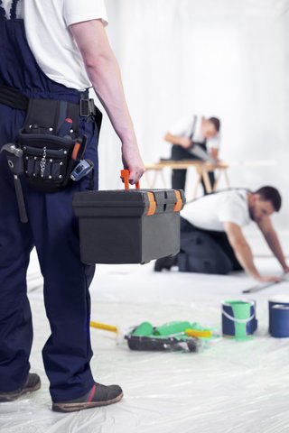 A construction worker holding a toolbox with two people working on a flooring project in the background.
