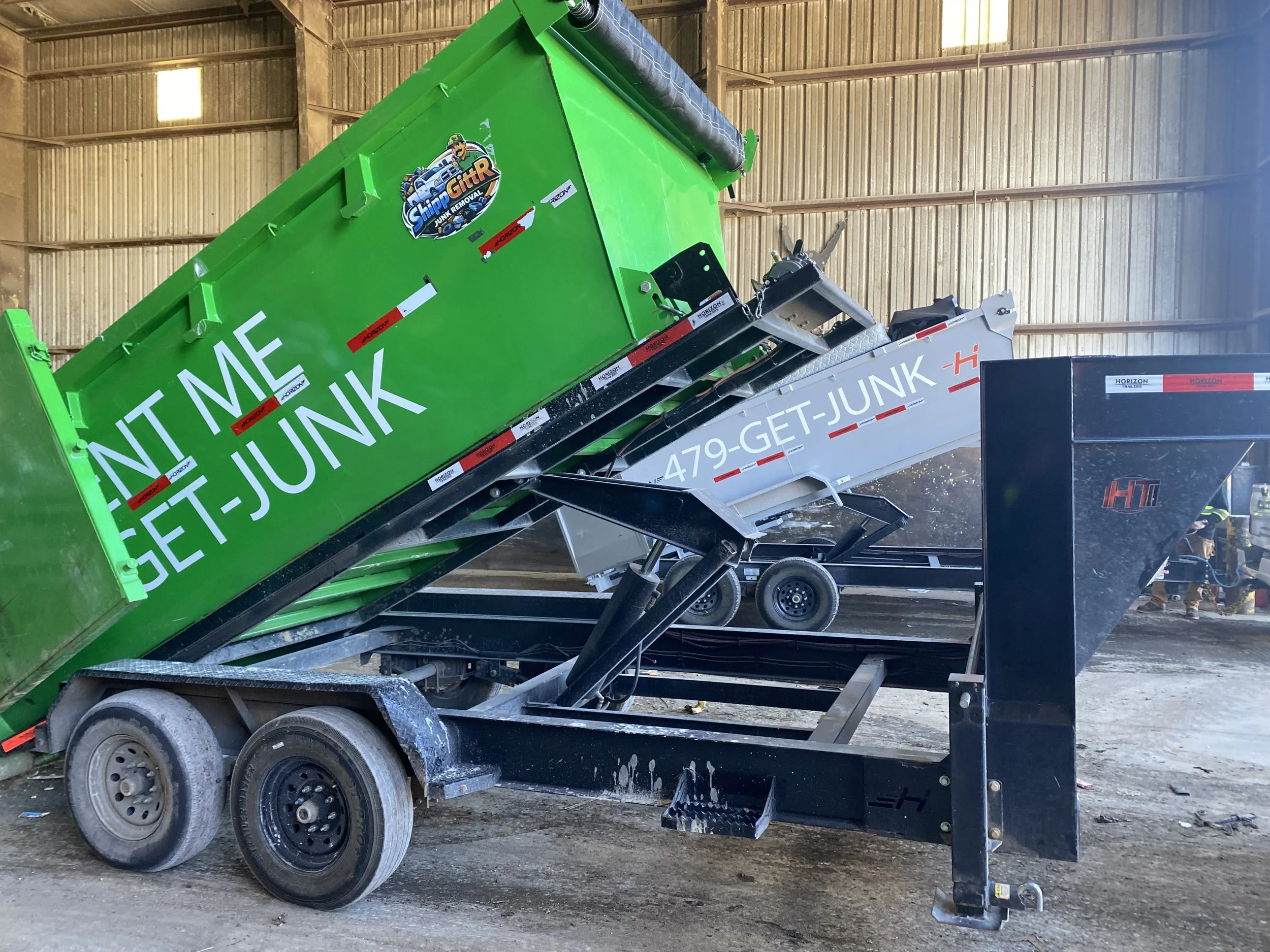 Two dump trailers, one green and one gray, inside a warehouse. The green trailer has a sticker with the words 'Ship GITR' and the phrase 'Come and get me, I'm junk' in white. The trailers are angled upward, showing their tipping mechanism.