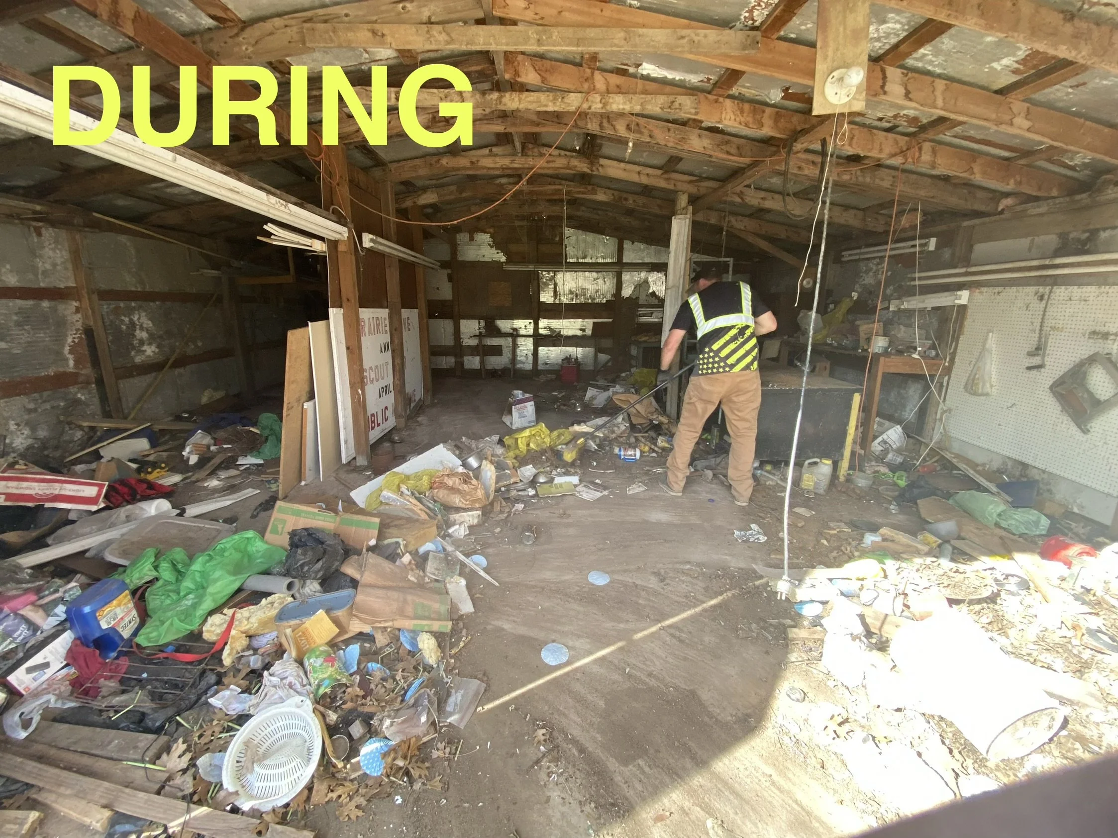 A person wearing a safety vest and khaki pants is cleaning up a cluttered garage or shed filled with trash, boxes, and debris. The garage has exposed wooden beams and a partially unfinished wall. The word 'DURING' is overlaid in large yellow letters at the top left of the image.