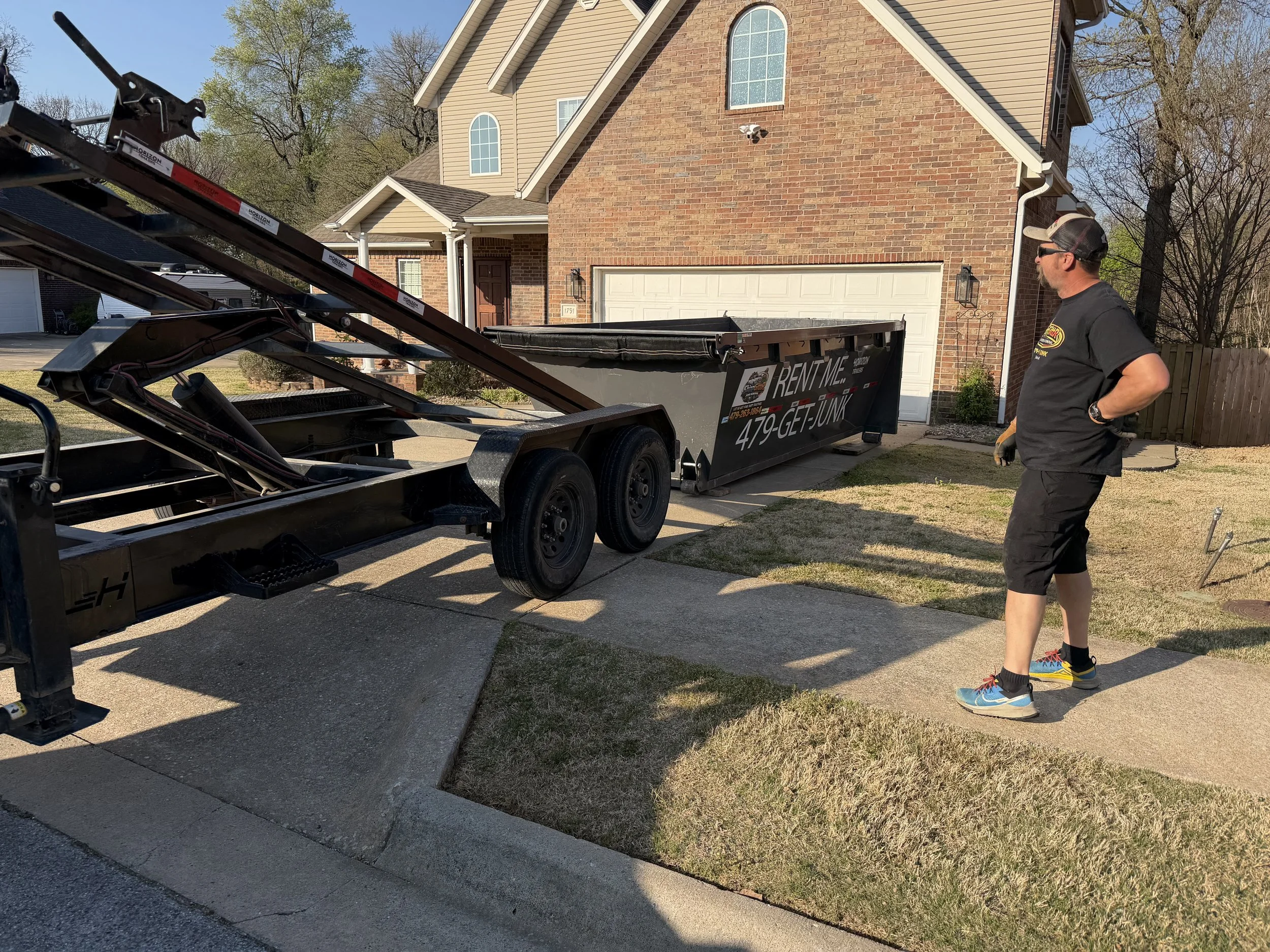 A man standing on the sidewalk observing a large flatbed trailer parked in front of a brick house. The trailer is tilted and has a black trash container with a sign offering rental services alongside a phone number.