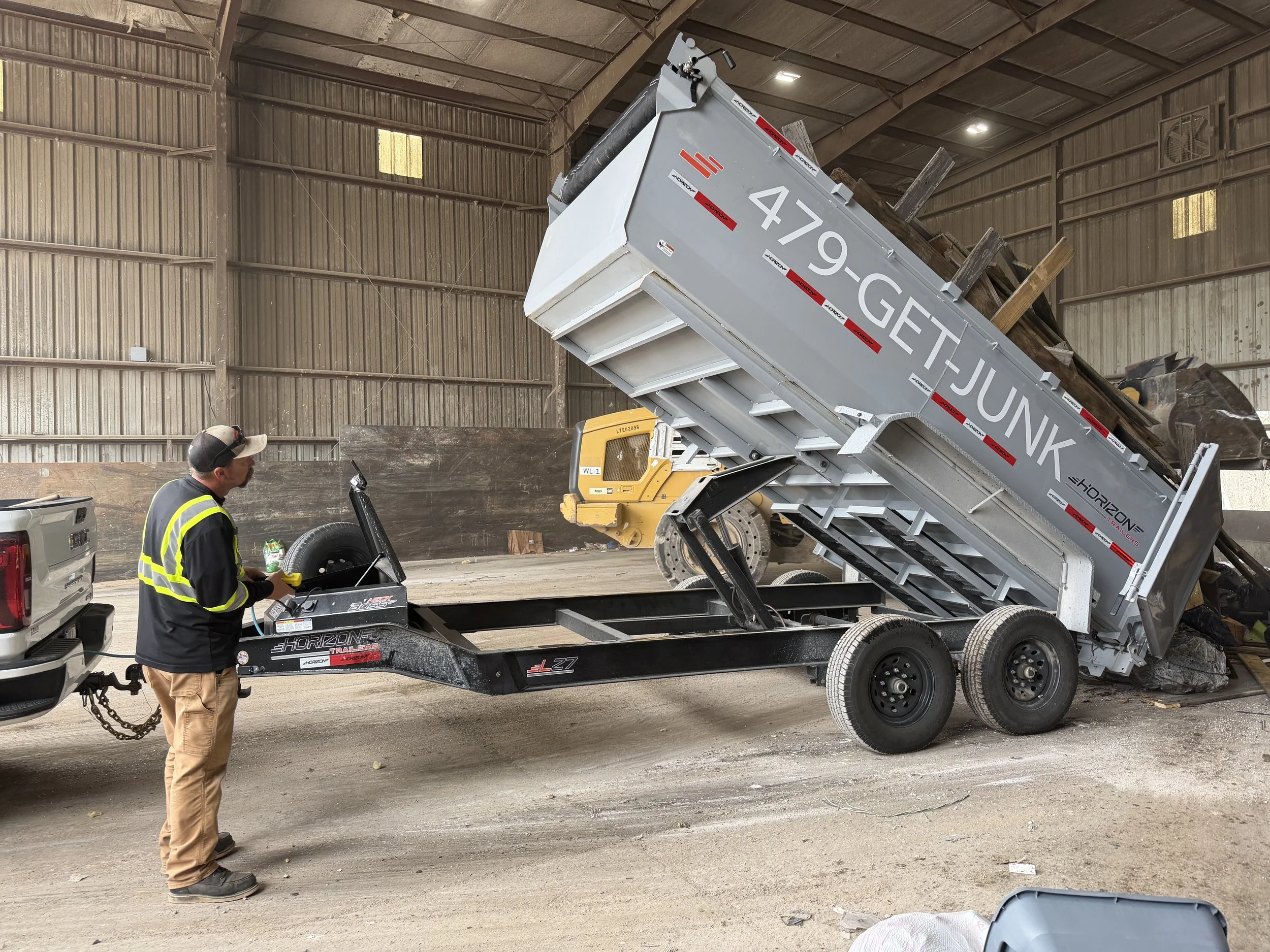 A person operating a yellow skid steer loader inside a warehouse, in front of a silver dump trailer filled with rocks or debris, which is tilted to unload its contents.