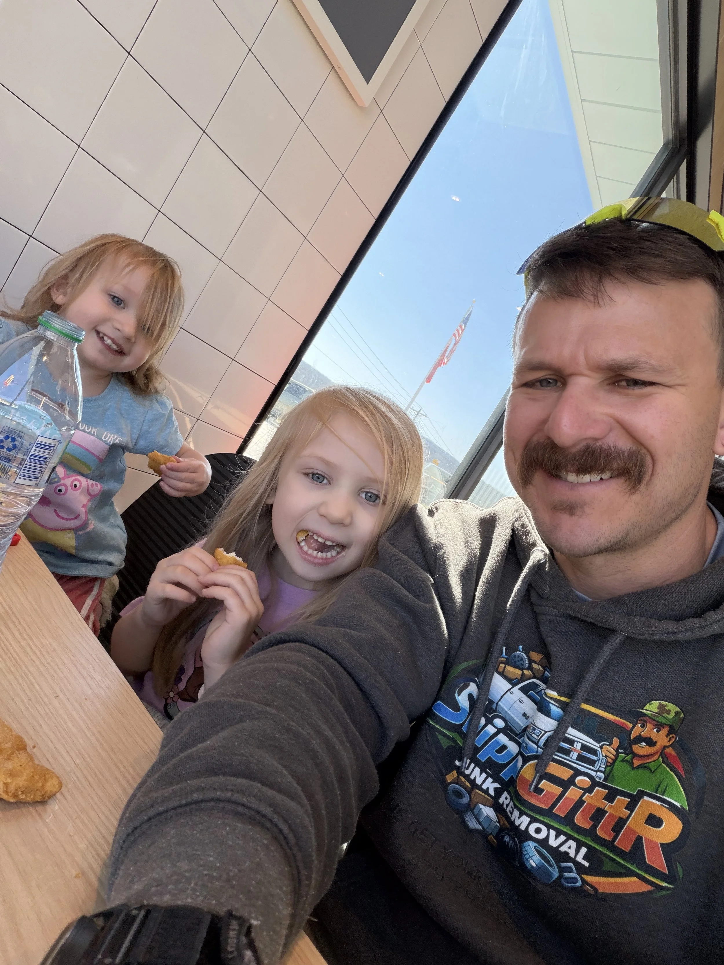 A man taking a selfie with two young girls in a restaurant. The man has a mustache and is wearing a dark hoodie with a logo. One girl has strawberry-blonde hair and is wearing a pink shirt, while the other girl has red hair and is wearing a Peppa Pig shirt. They are smiling and eating chicken nuggets.