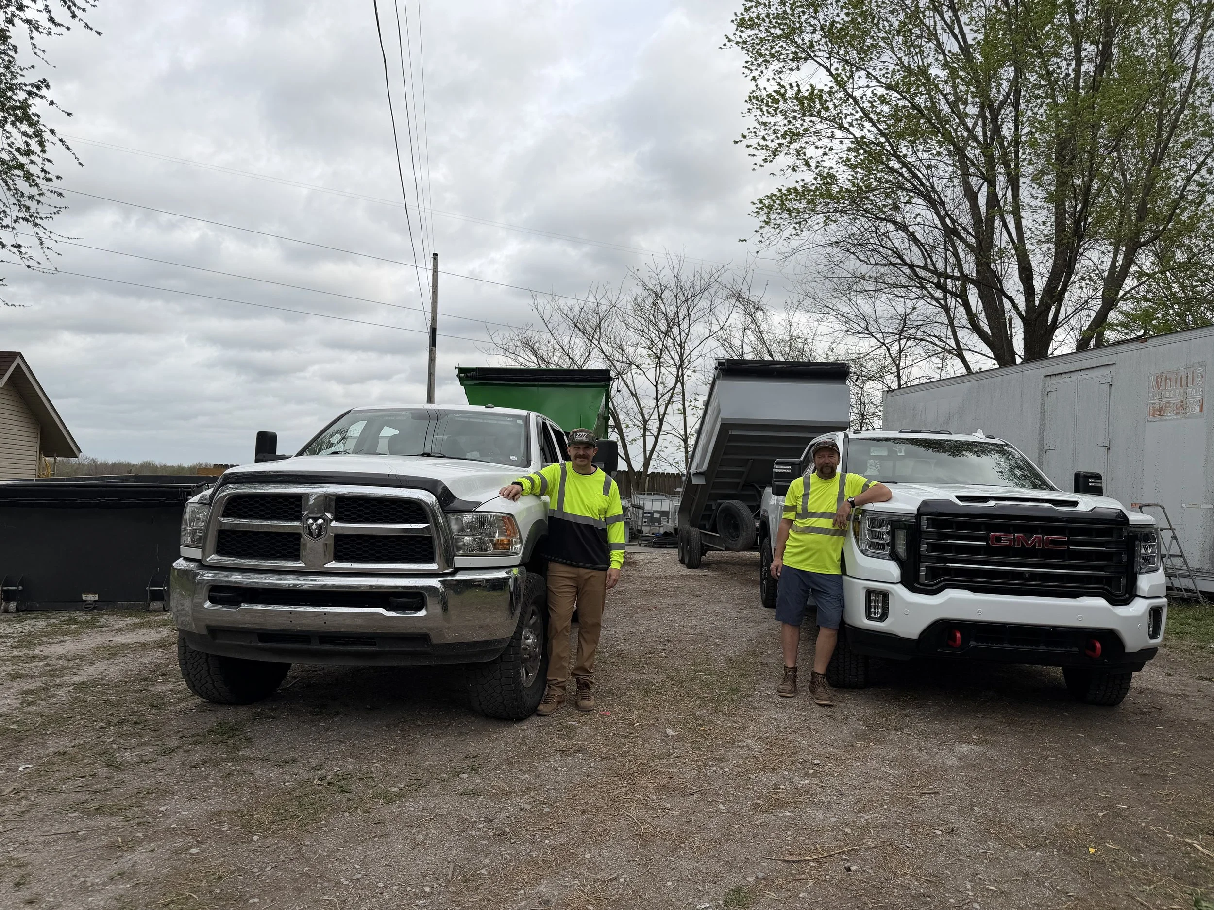 Two men wearing safety vests standing next to white trucks with dump beds parked on gravel, in an outdoor area with trees and portable containers in the background.