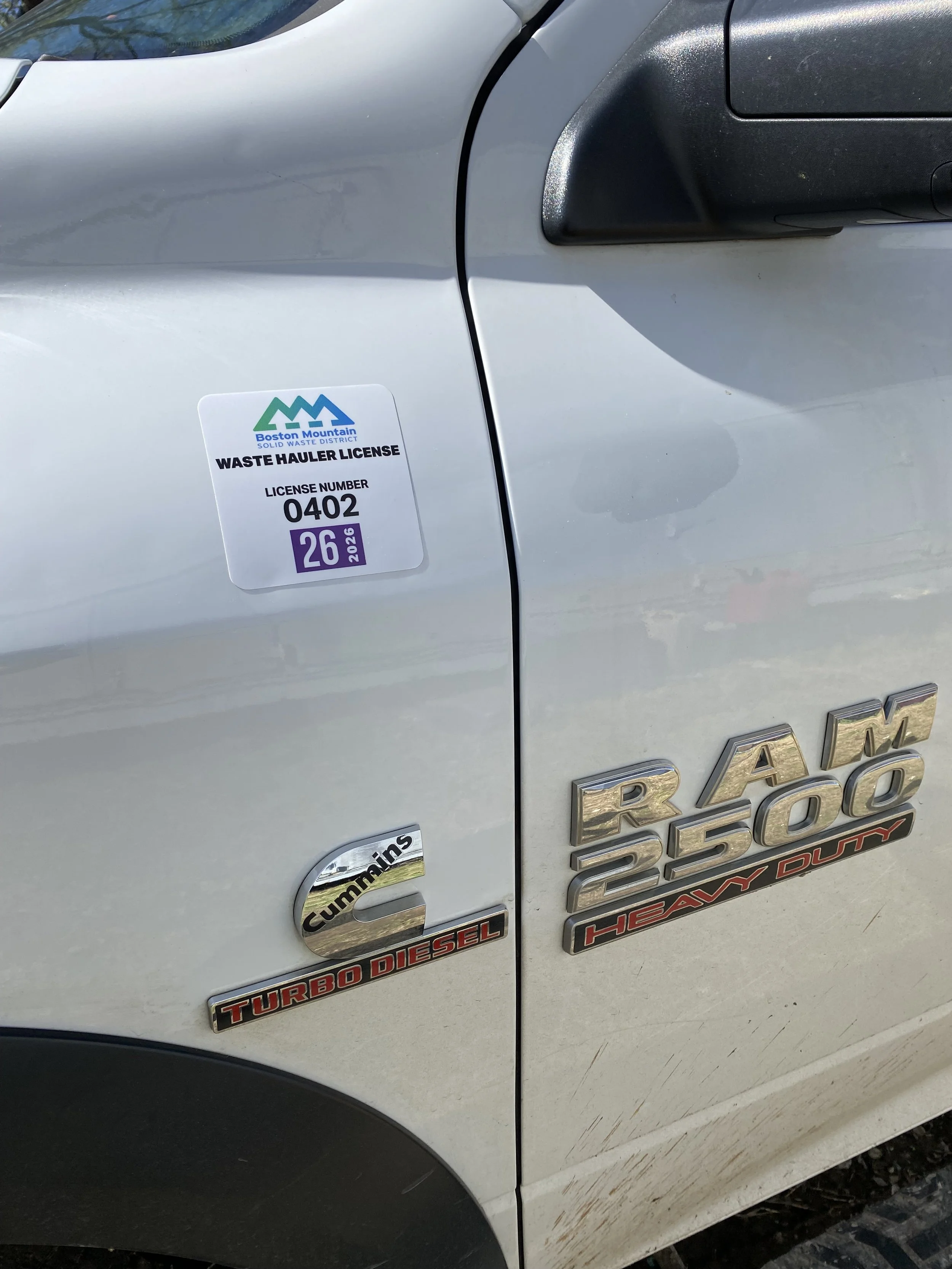 Close-up of a white RAM 2500 heavy-duty pickup truck with badges for Cummins Turbo Diesel and Heavy Duty, and a Boston Mountain Waste District waste hauler license sticker.