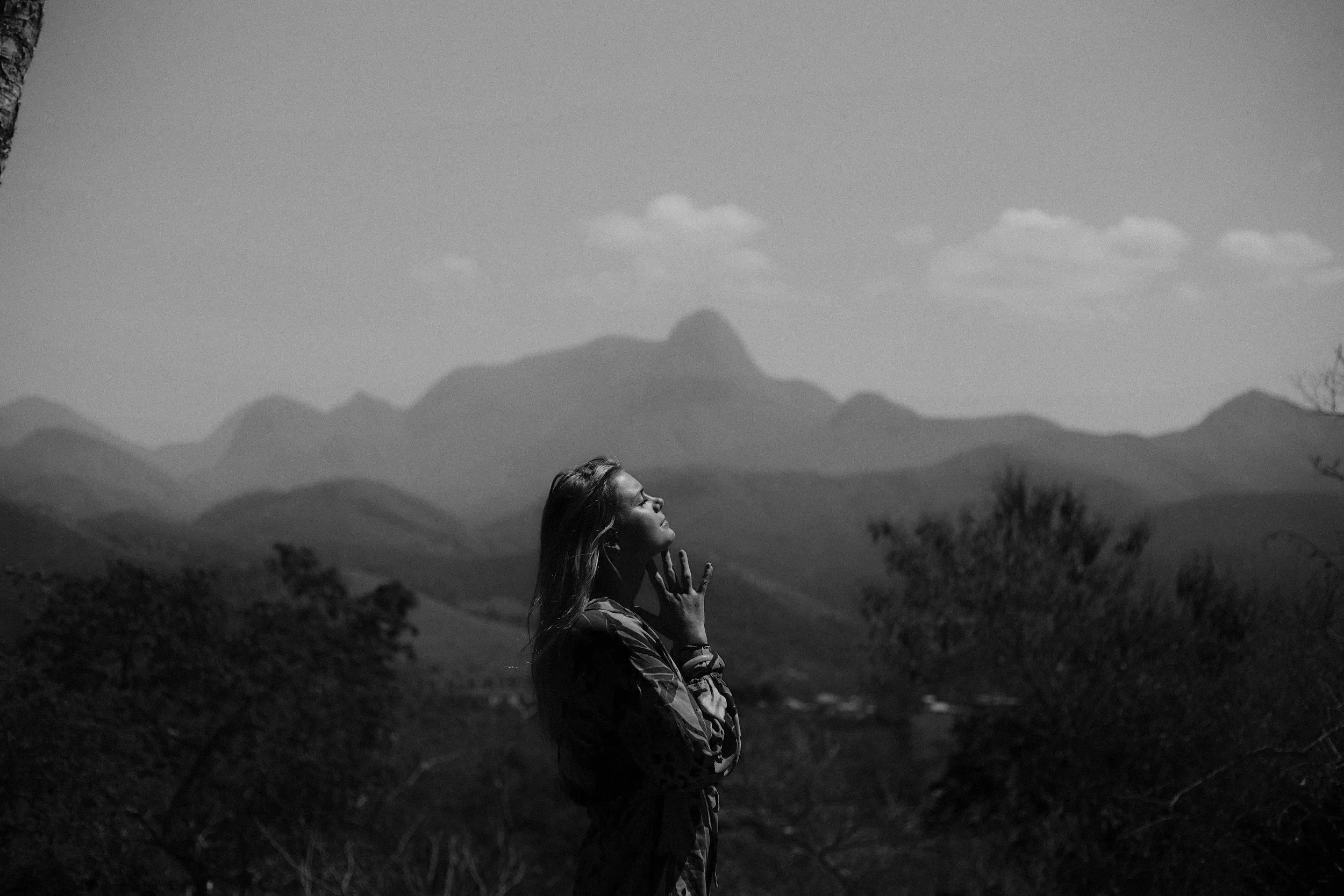 A woman standing outdoors with her eyes closed and hand on her cheek, surrounded by trees and mountains in the background, in black and white.
