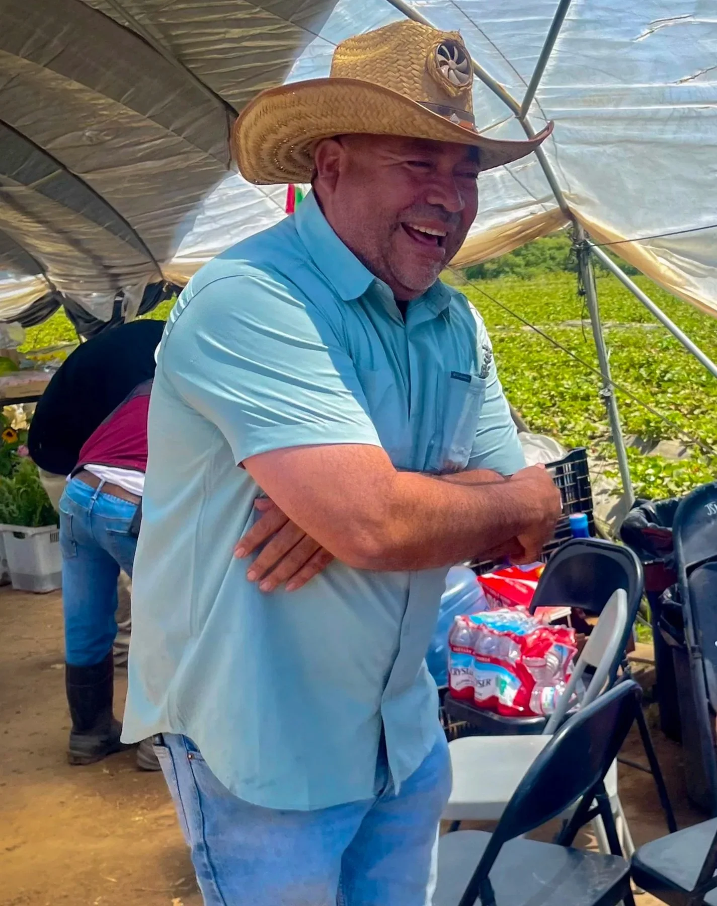 A man wearing a large straw cowboy hat and a light blue shirt, standing inside a greenhouse or tent, smiling and with arms crossed.