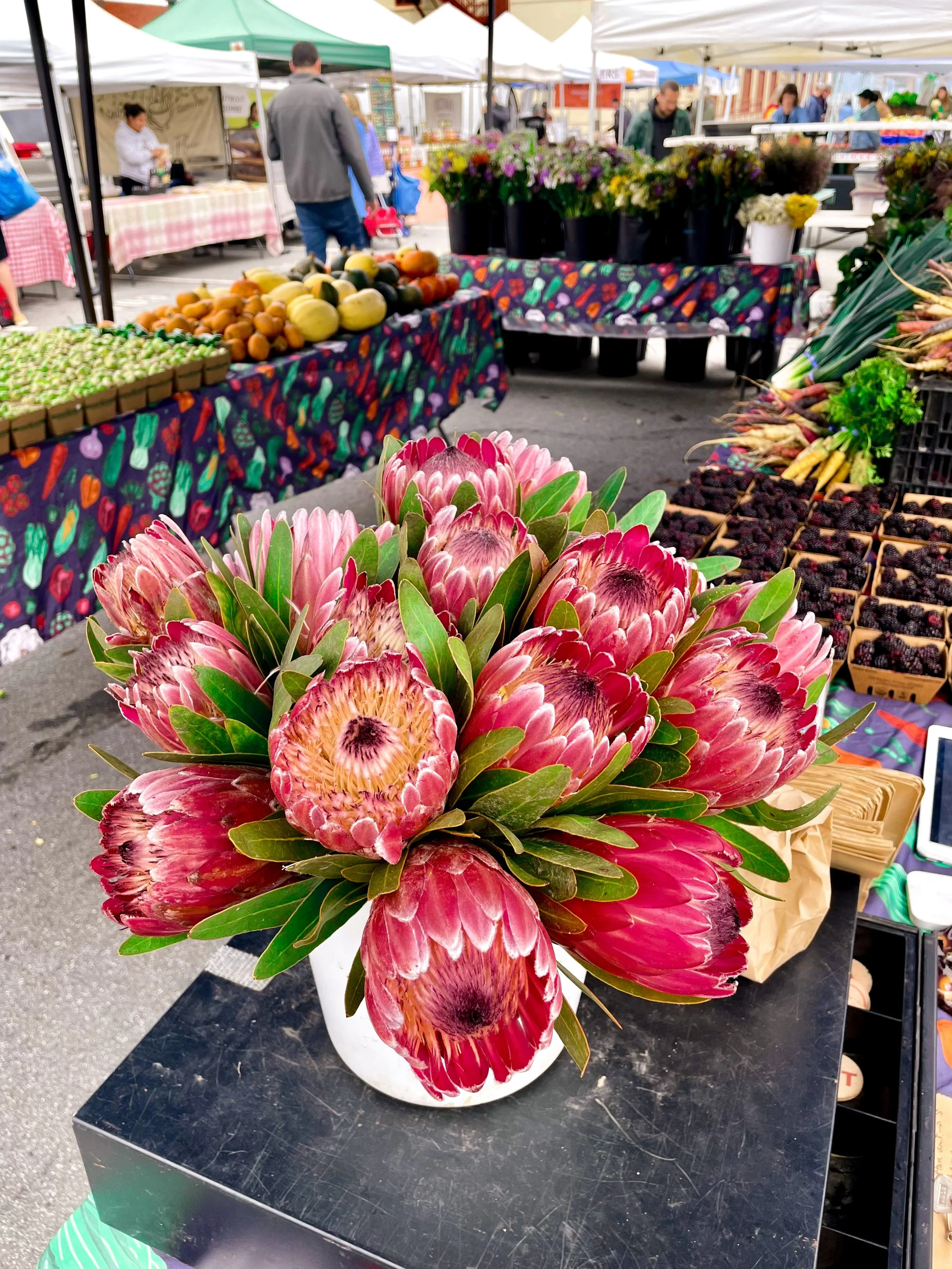 A bouquet of pink protea flowers in a white vase at an outdoor farmers market with produce and vendors in the background.
