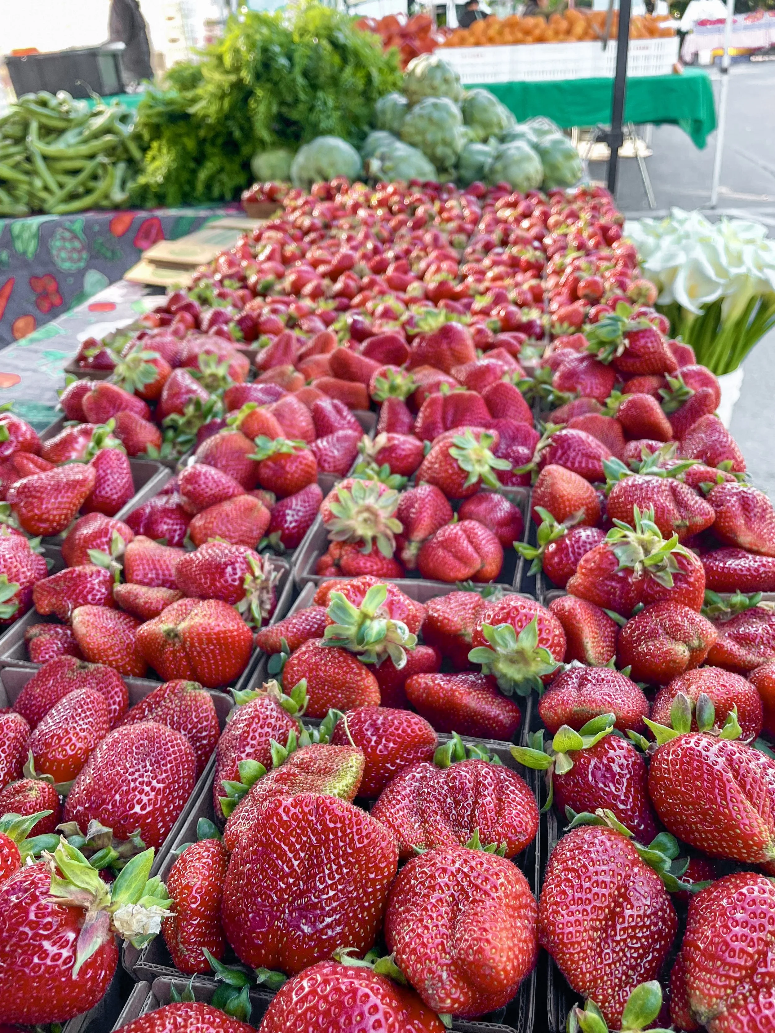 Fresh strawberries displayed at an outdoor market stall with other vegetables and fruits in the background.