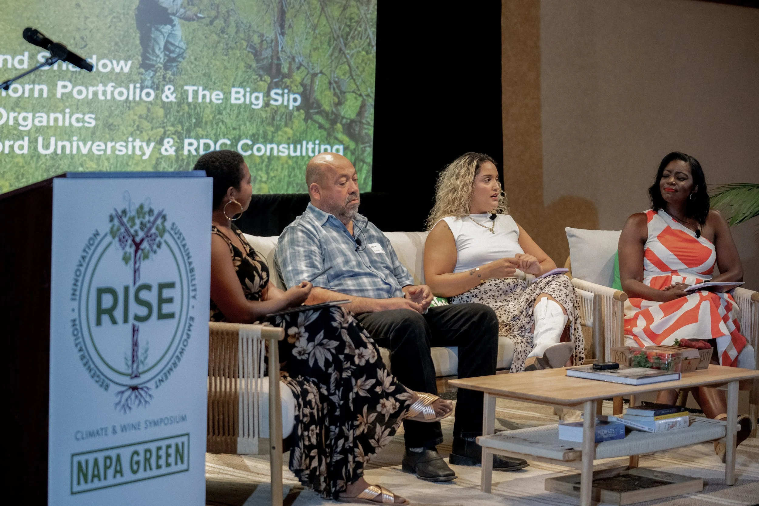 A panel of four people seated on a white sofa at a conference, with a screen behind them displaying event information and the Napa Green logo on a stand to the left. The scene appears to be a climate and wine symposium.