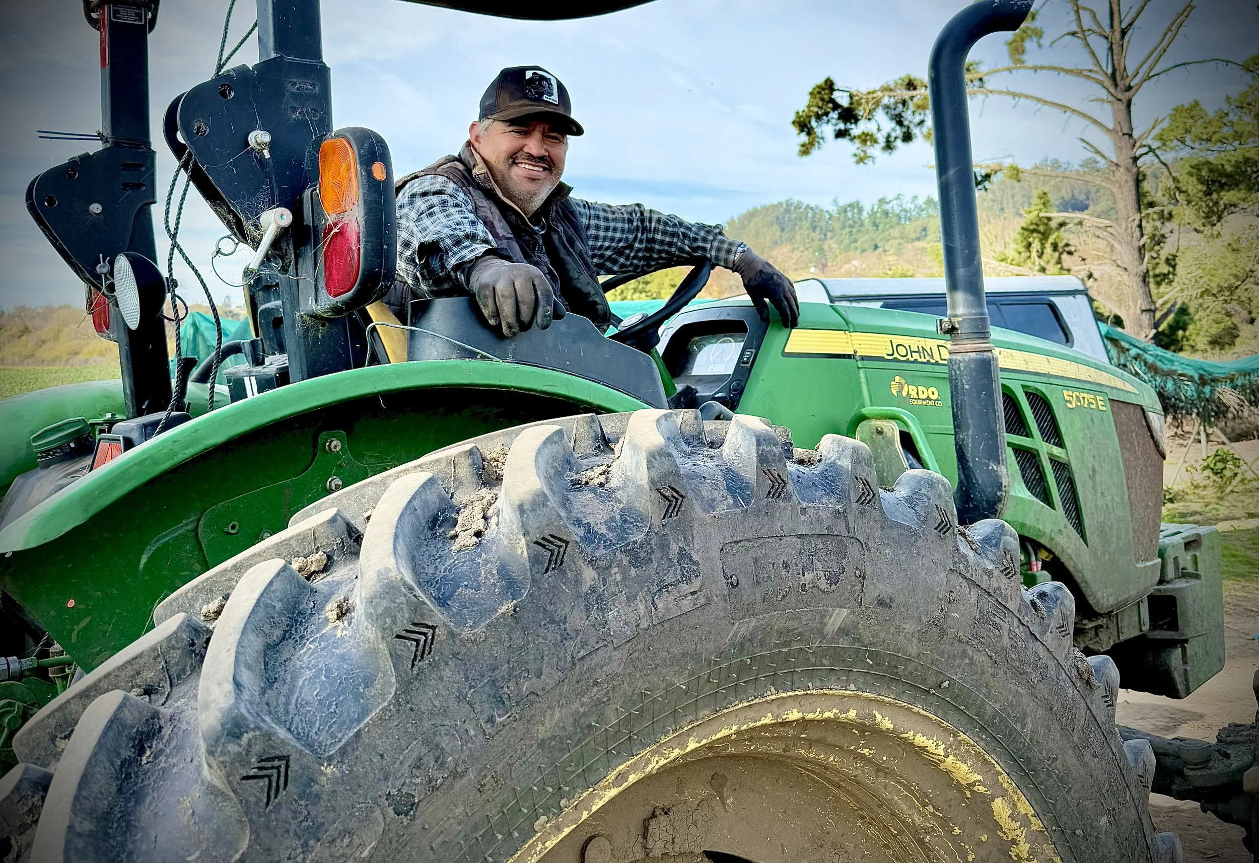 A man wearing a plaid shirt, black vest, and a baseball cap sitting on a green tractor, smiling at the camera with trees and a blue sky in the background.
