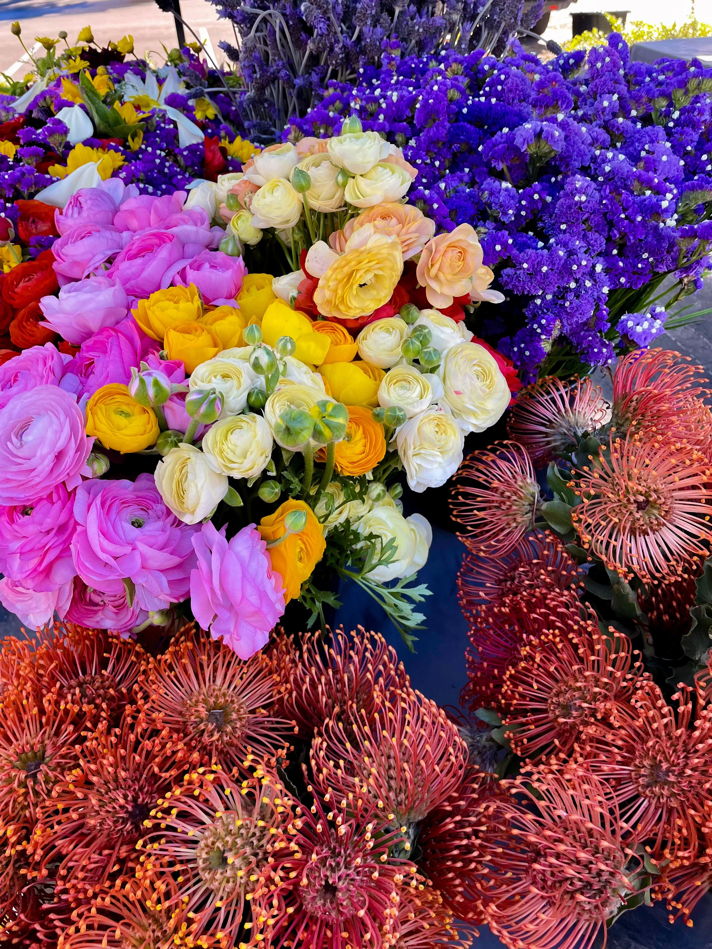 A colorful display of various flowers, including pink ranunculus, yellow and white ranunculus, purple statice, and red pincushion protea.