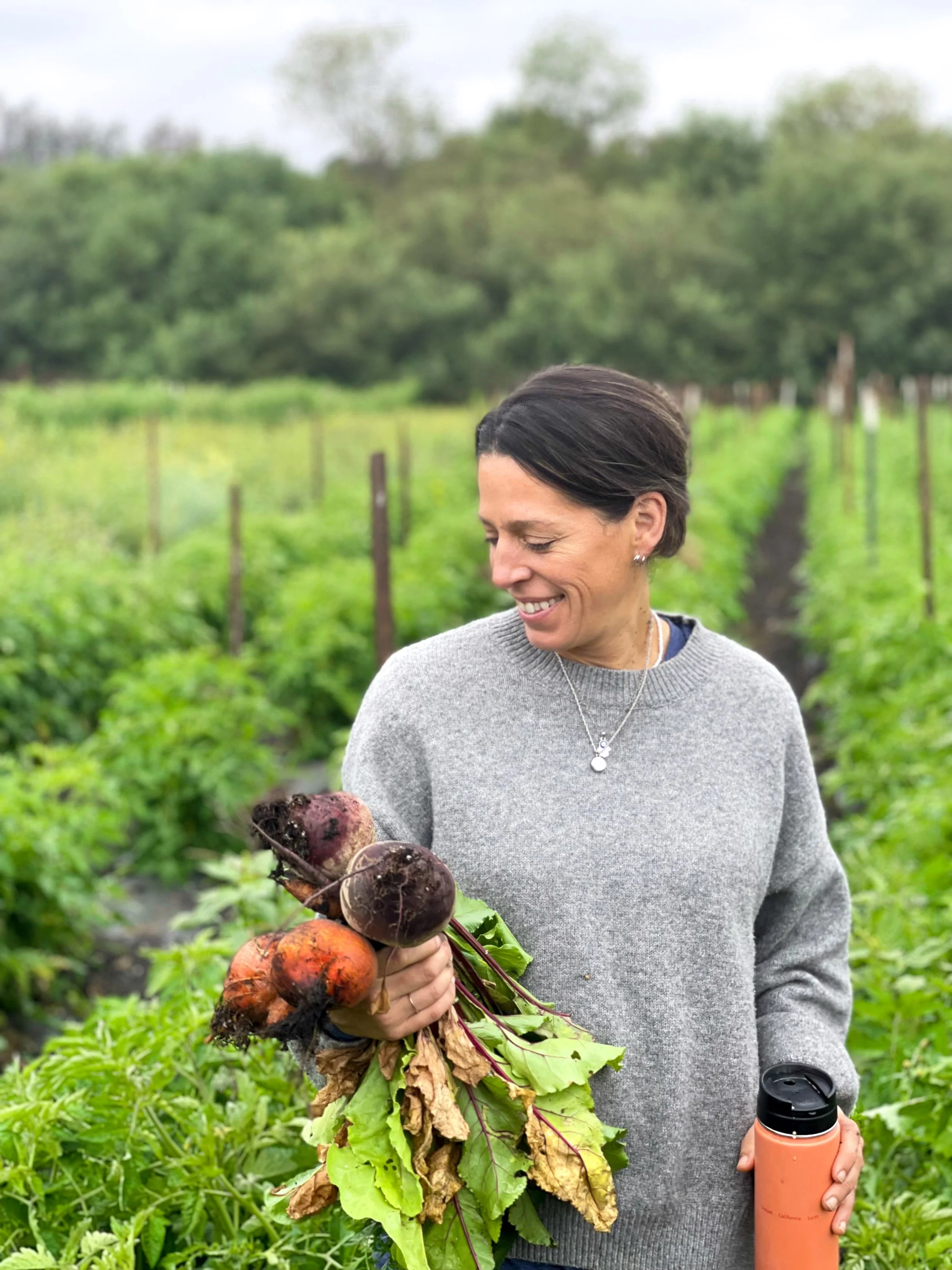 A woman holding freshly picked beets and carrots in a vegetable garden, smiling, next to a travel mug, with green plants and trees in the background.