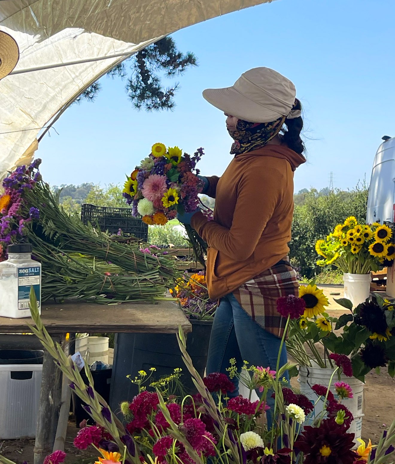 A woman at an outdoor flower stand sorting a bouquet of colorful flowers, wearing a wide-brimmed hat, a face mask, a brown jacket, and jeans. The stand is filled with various flower arrangements and loose flowers, with a tree and blue sky in the back