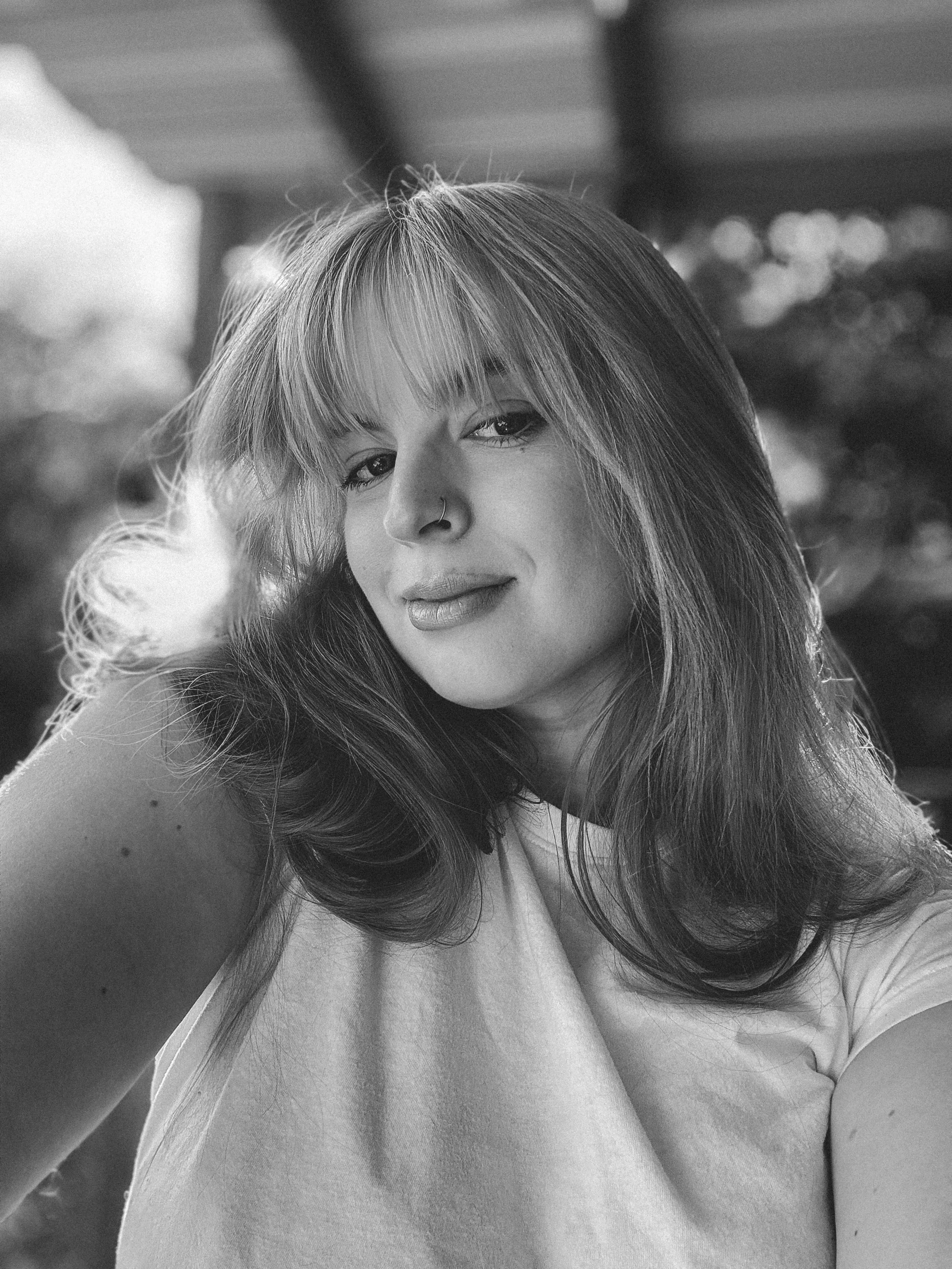 A black-and-white portrait of a young woman with long hair, wearing a nose ring, and looking at the camera with a neutral expression.