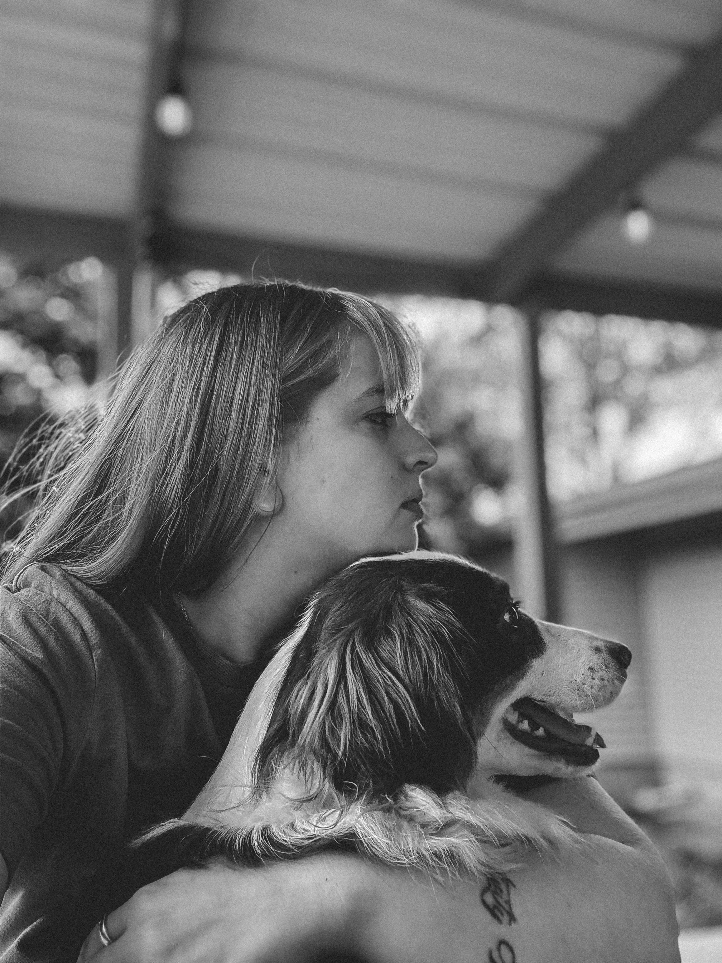 A woman with long hair and a tattoo on her arm is holding a dog with a black and white face, the woman is looking to the side and smiling.