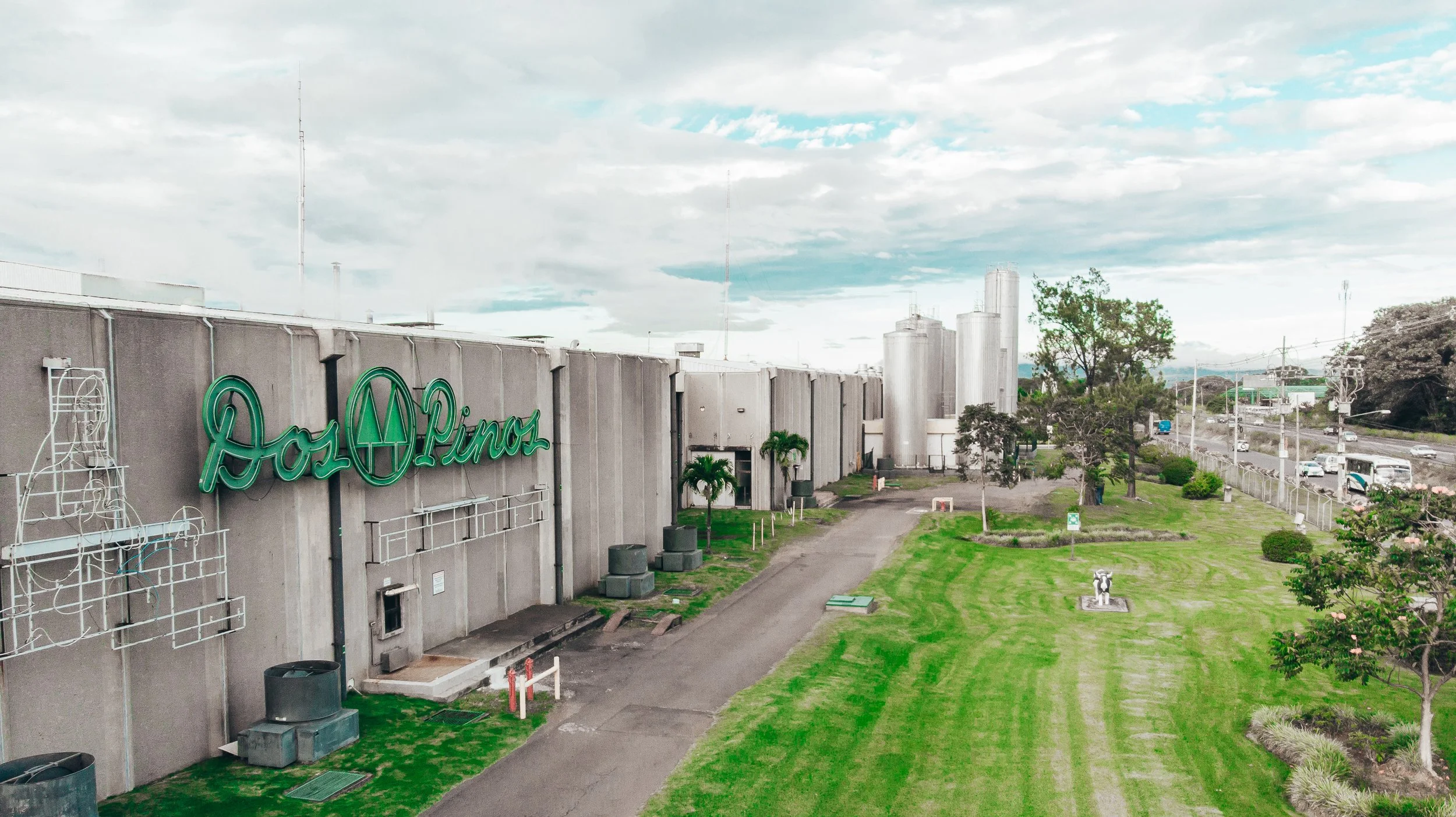 Vista de un edificio con el cartel de 'Los Pinos' en verde y un área verde con árboles y caminos en primer plano, al fondo un cielo nublado y una ciudad.