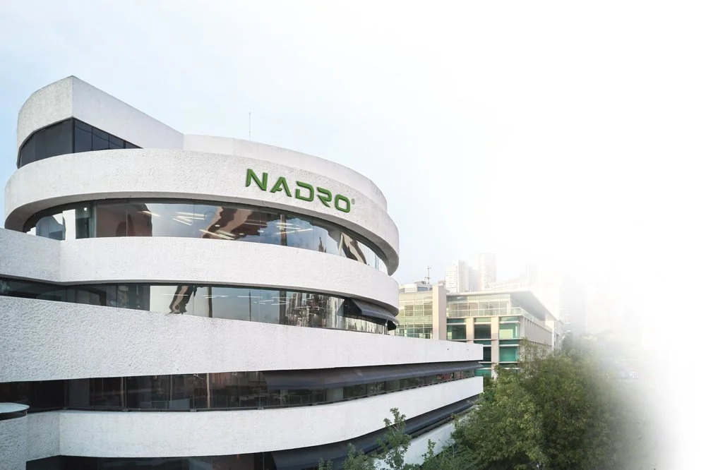 Edificio moderno de forma curva con ventanas de cristal y señal que dice 'NADRO' en la parte superior, en un entorno urbano con otros edificios y árboles.